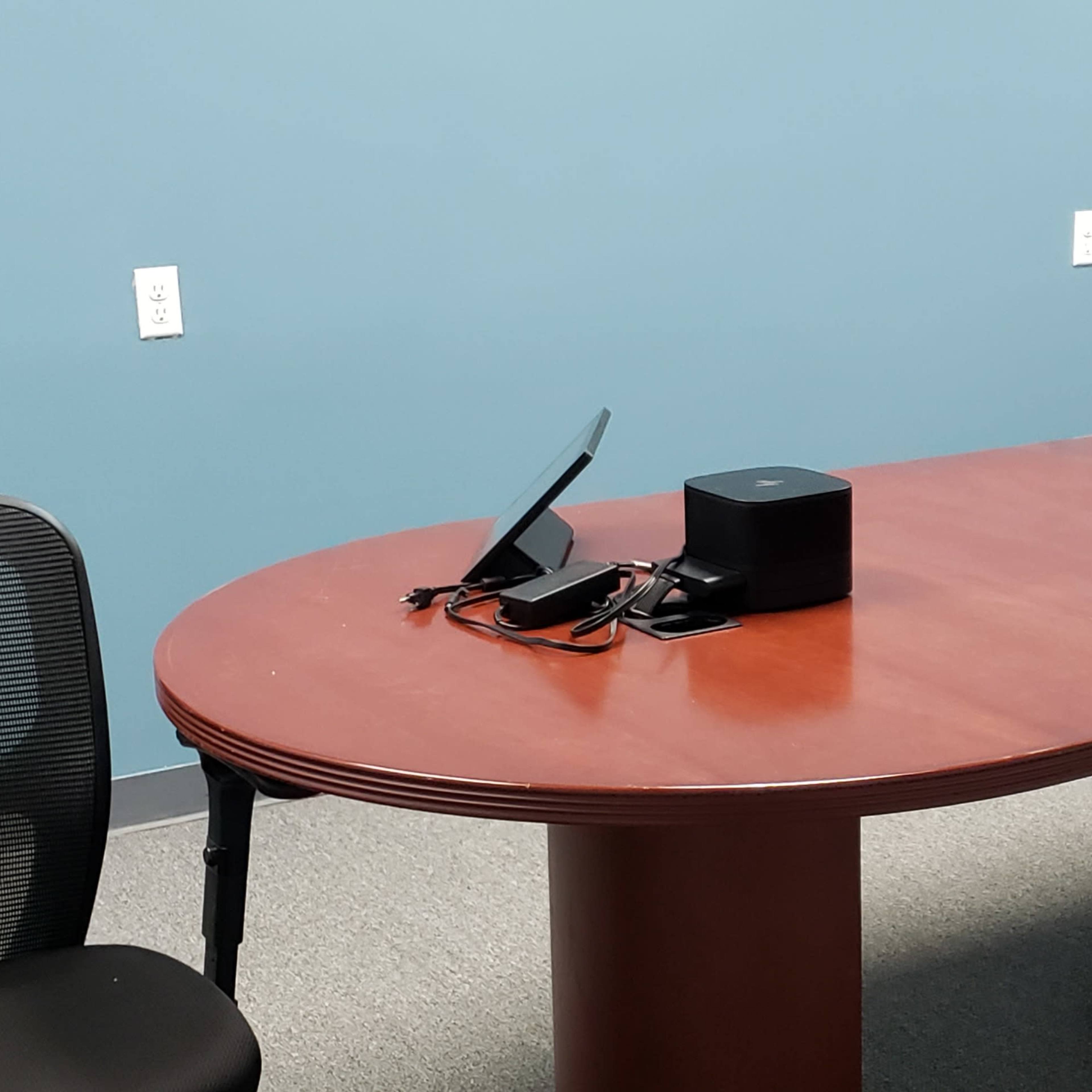 A large wooden conference table is positioned in a room with blue walls, accompanied by a black chair and a small electronic device on the table.