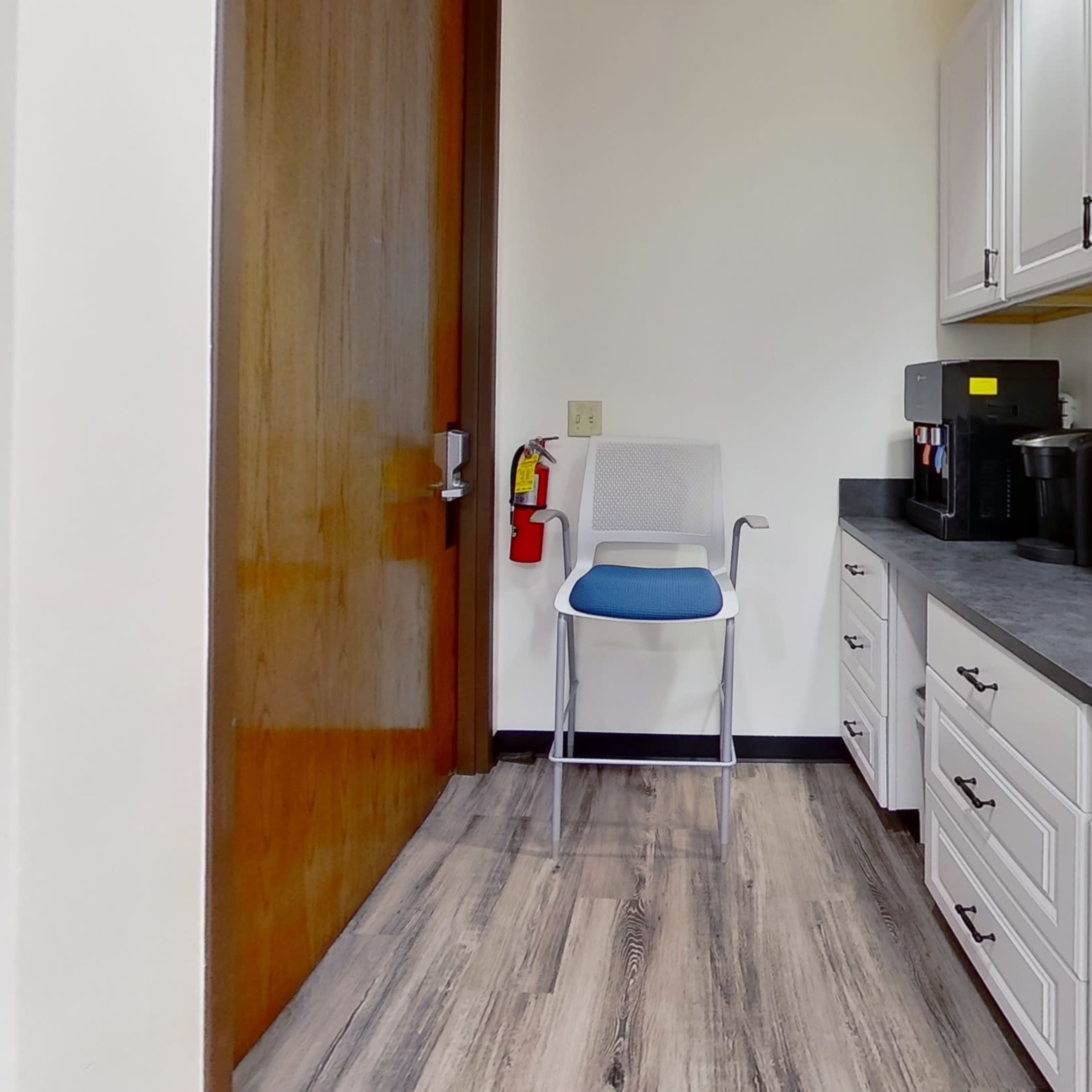 A small kitchen area features white cabinets, a blue chair, and a coffee machine along a dark countertop.