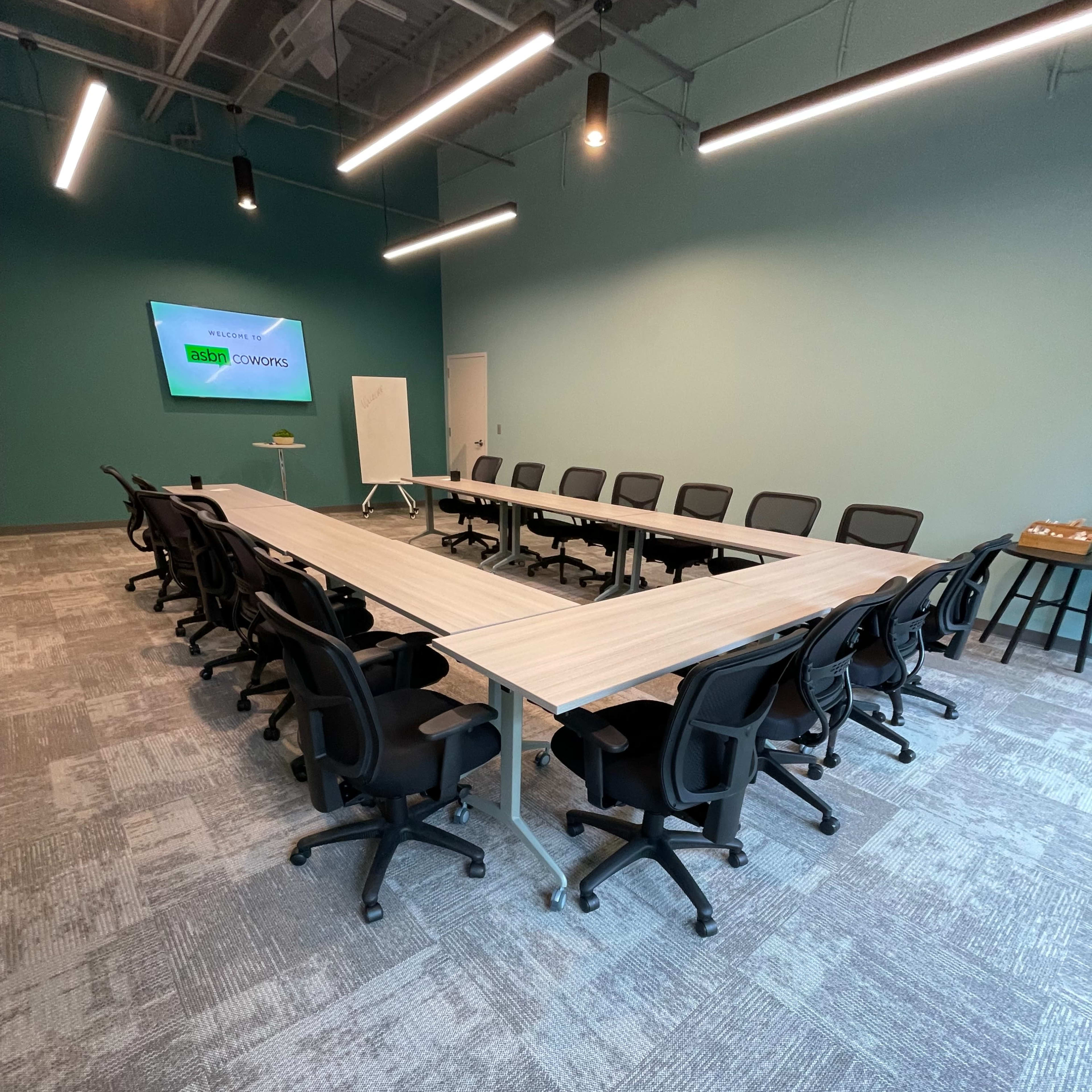 A rectangular conference room features a long table surrounded by black office chairs, with a large screen mounted on a light blue wall.