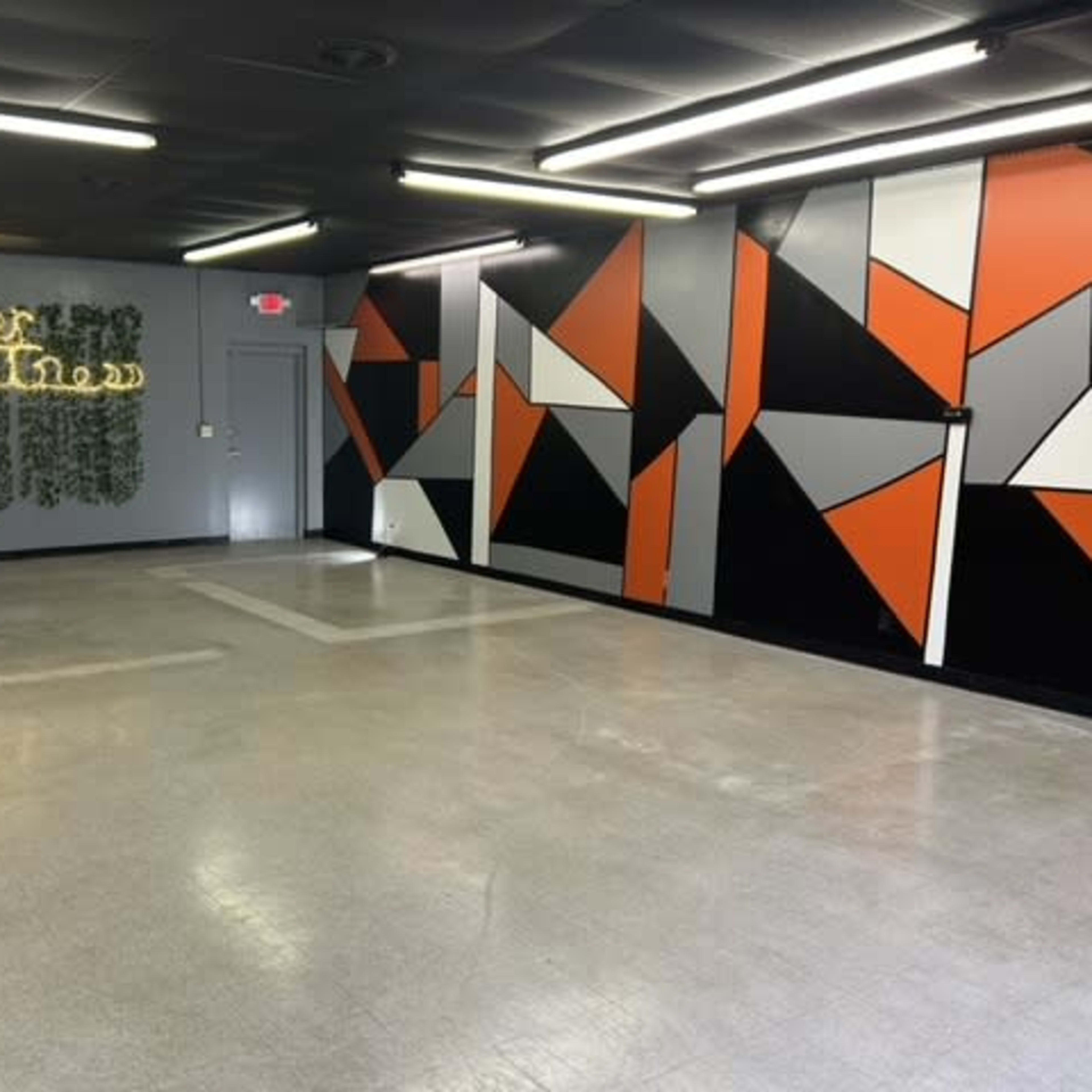 A spacious room with a concrete floor, featuring painted geometric designs in black, orange, and white on one wall, and neon lights with the words "Not the Same" on another wall.