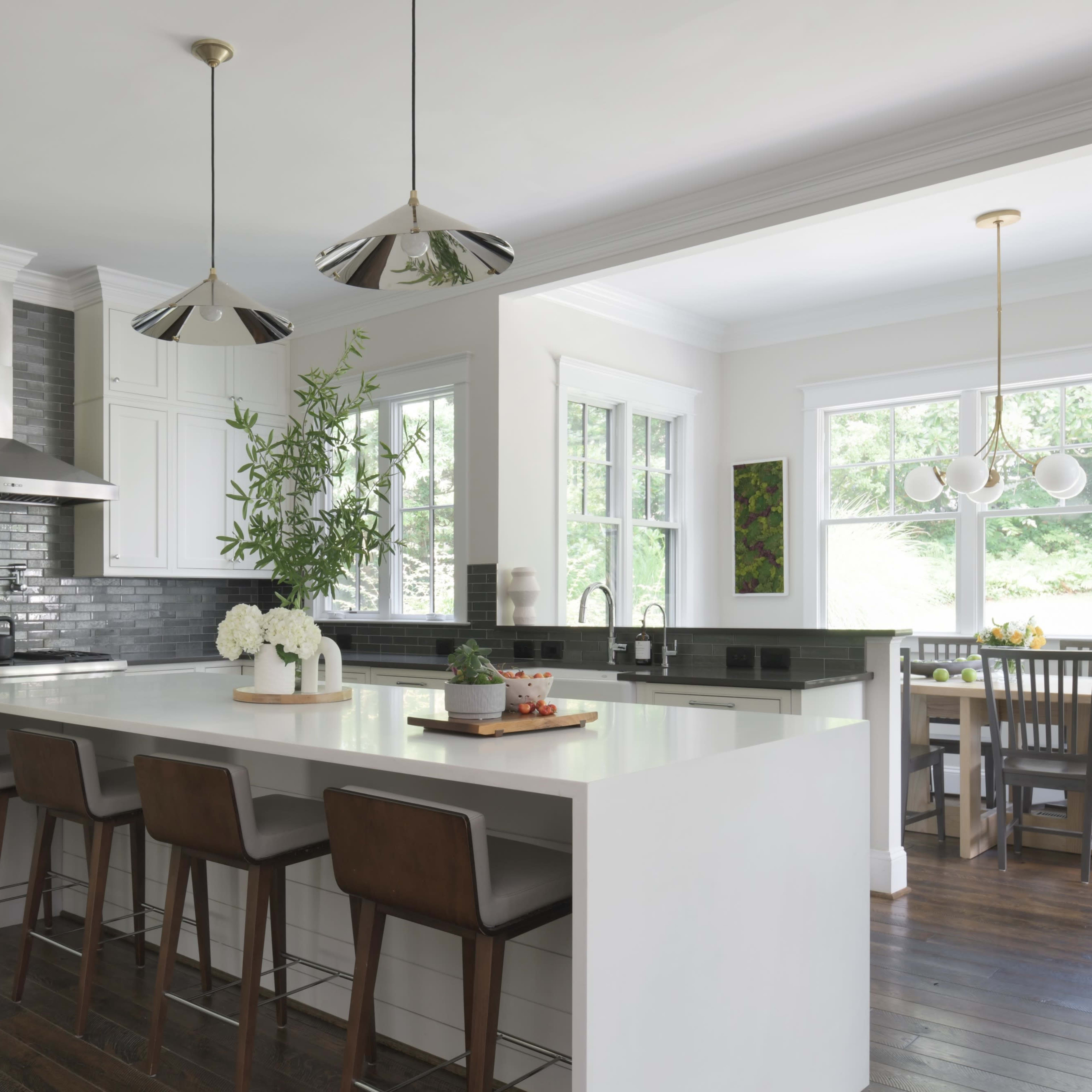 A modern kitchen features a large white island with four brown stools, stainless steel appliances, and natural light streaming through the windows.