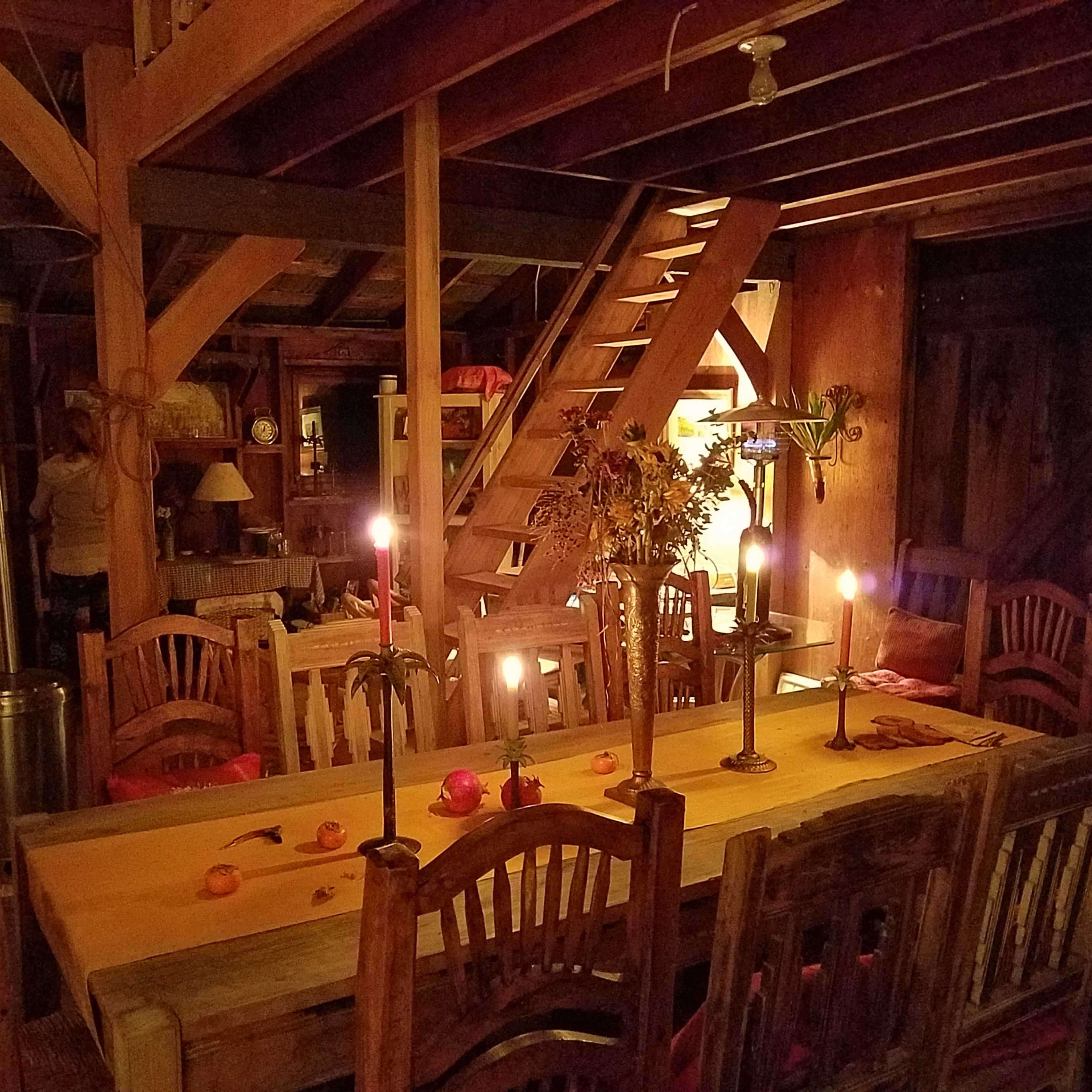 A warmly lit wooden dining area with a long table surrounded by chairs, a staircase in the background, and candles providing soft illumination.