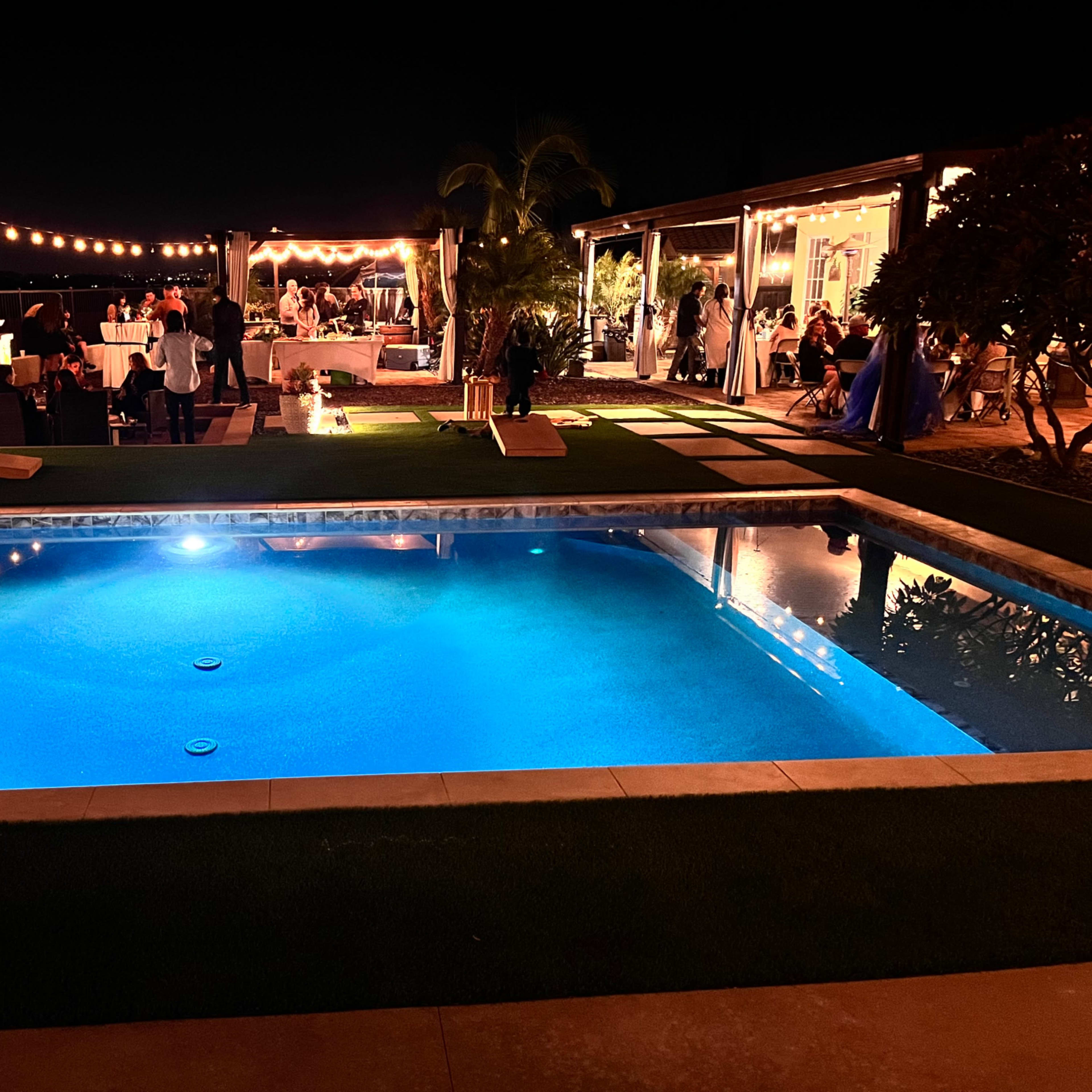 A brightly lit pool area is set for a nighttime celebration with string lights overhead and guests mingling by a decorated outdoor space.