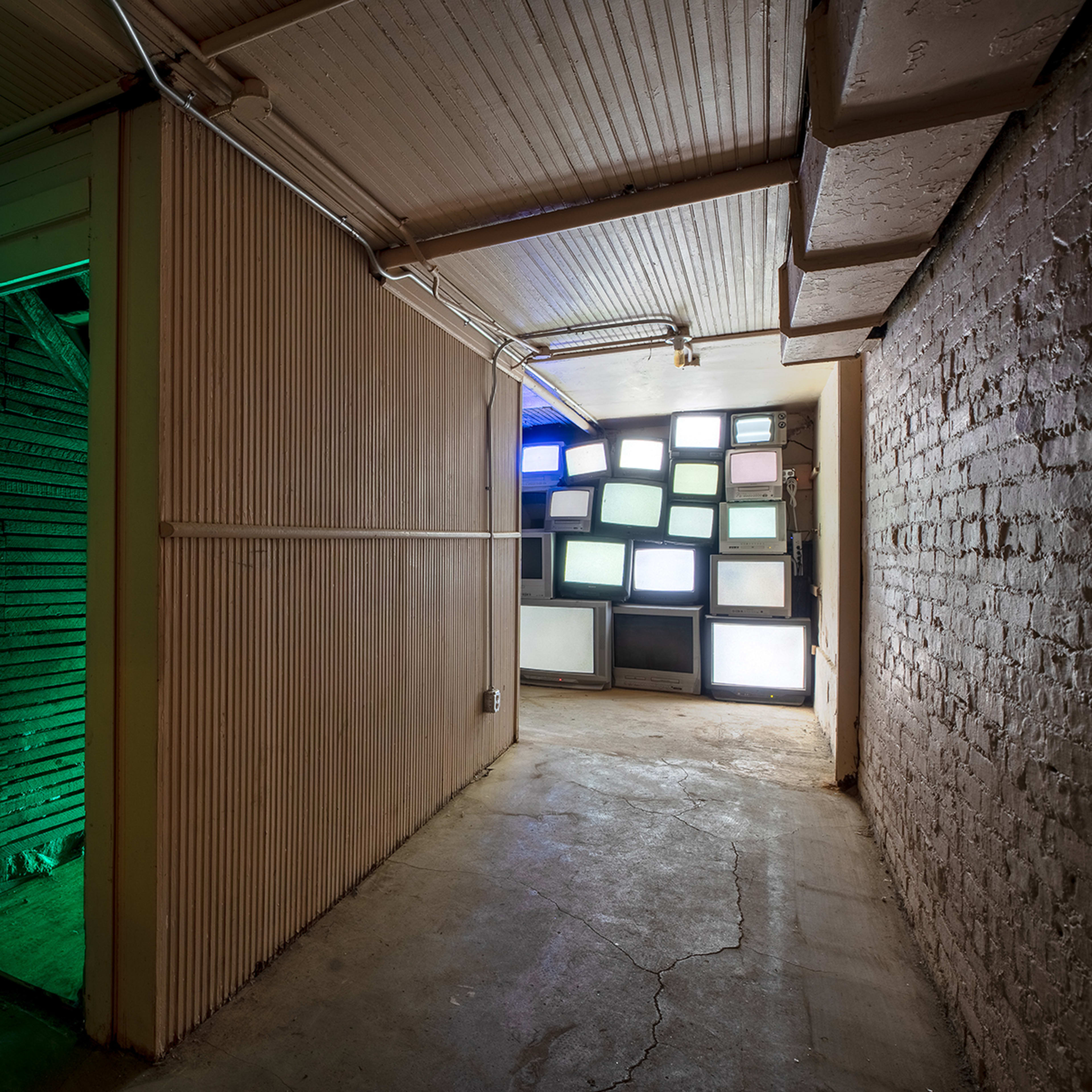 A narrow hallway leads to a wall of illuminated television screens, with a green-lit doorway on the left.