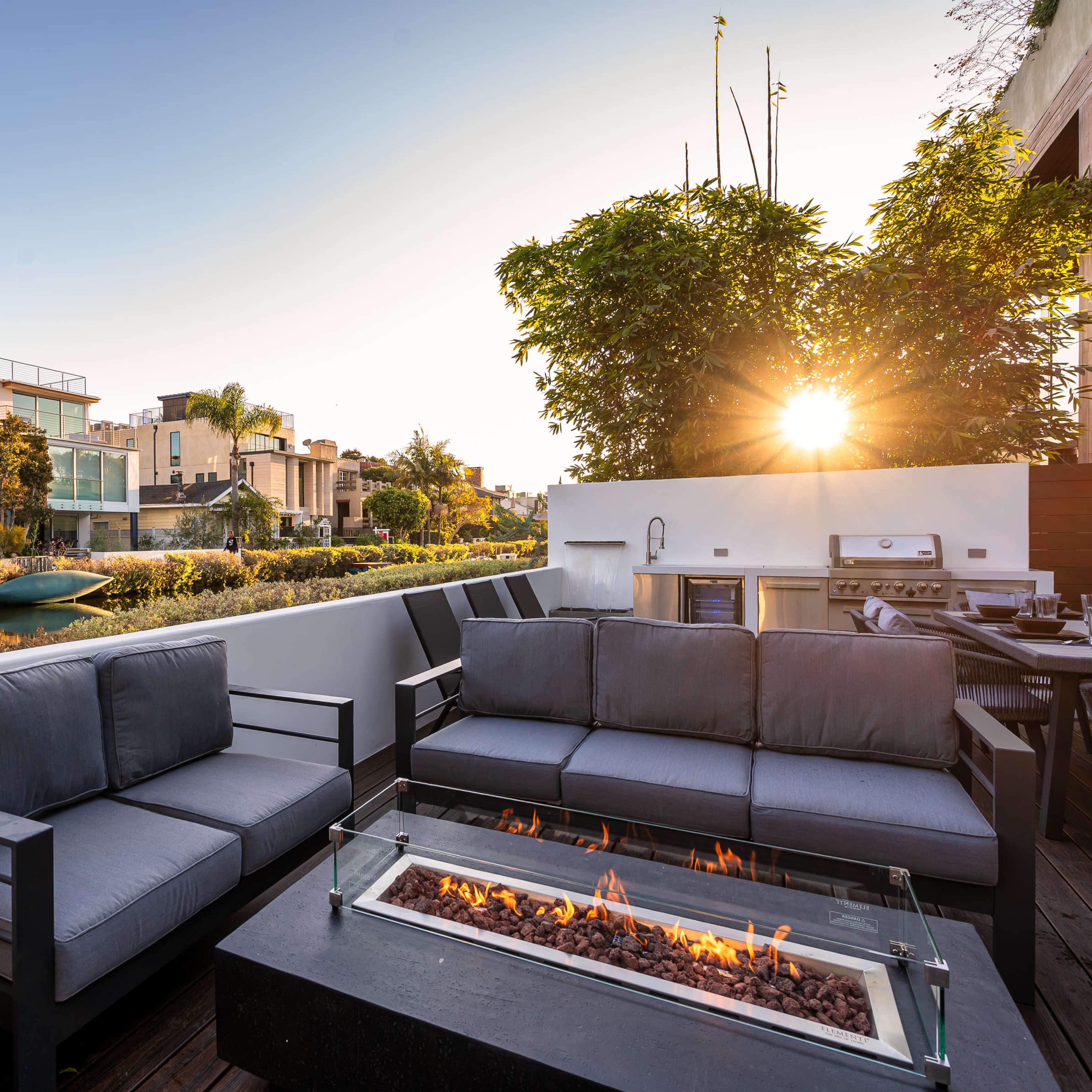A modern outdoor seating area features gray couches surrounding a glass fire pit, with a sunset illuminating a canal lined by residential buildings.