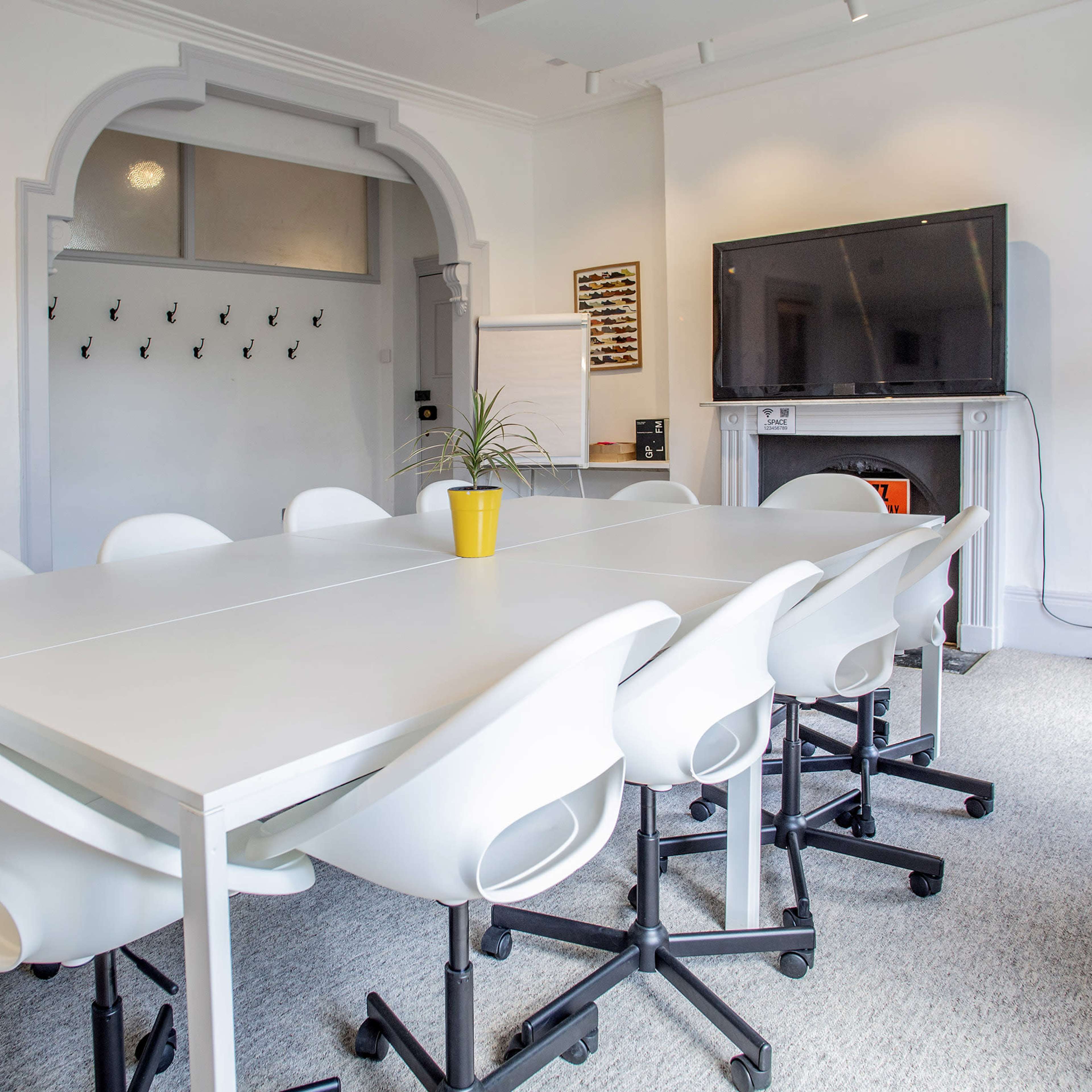 A bright conference room with a large white table surrounded by white chairs, a television on the wall, and a plant on a side table.