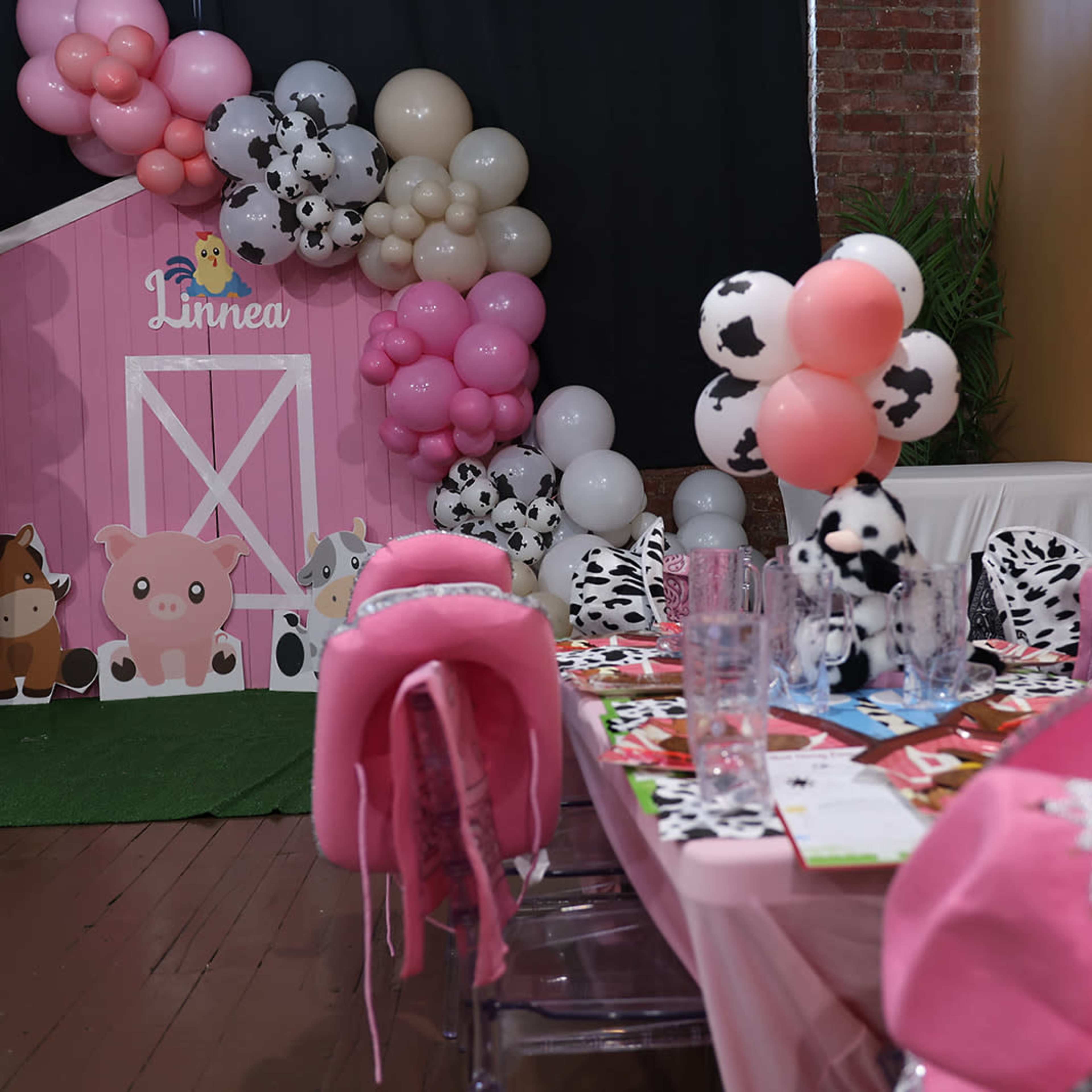 A decorated party space features a pink barn backdrop, colorful balloons, and a table set with cow-themed decorations and seating.