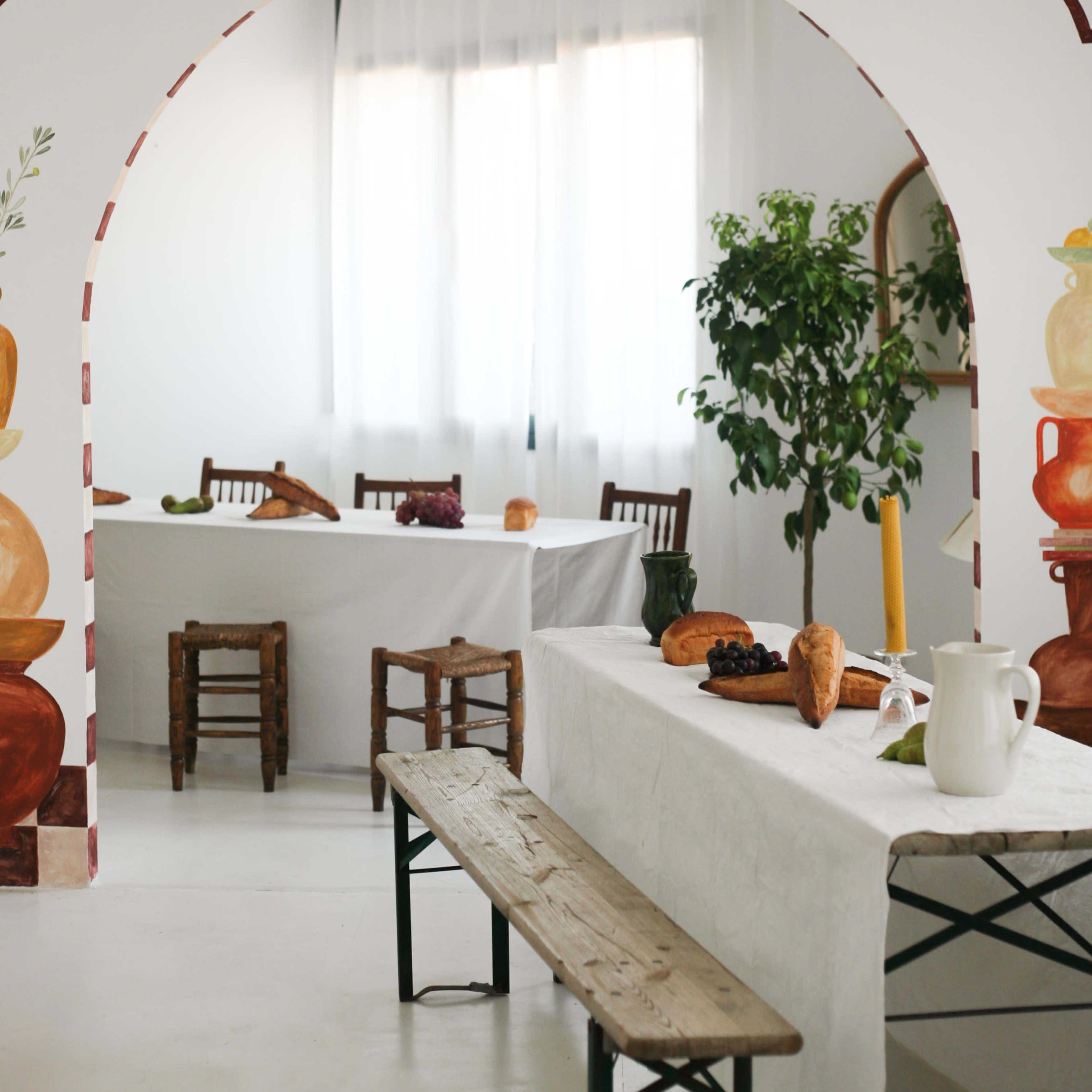 A bright, minimalistic dining space features a large archway with painted celestial designs, leading to tables adorned with white tablecloths and simple decor.