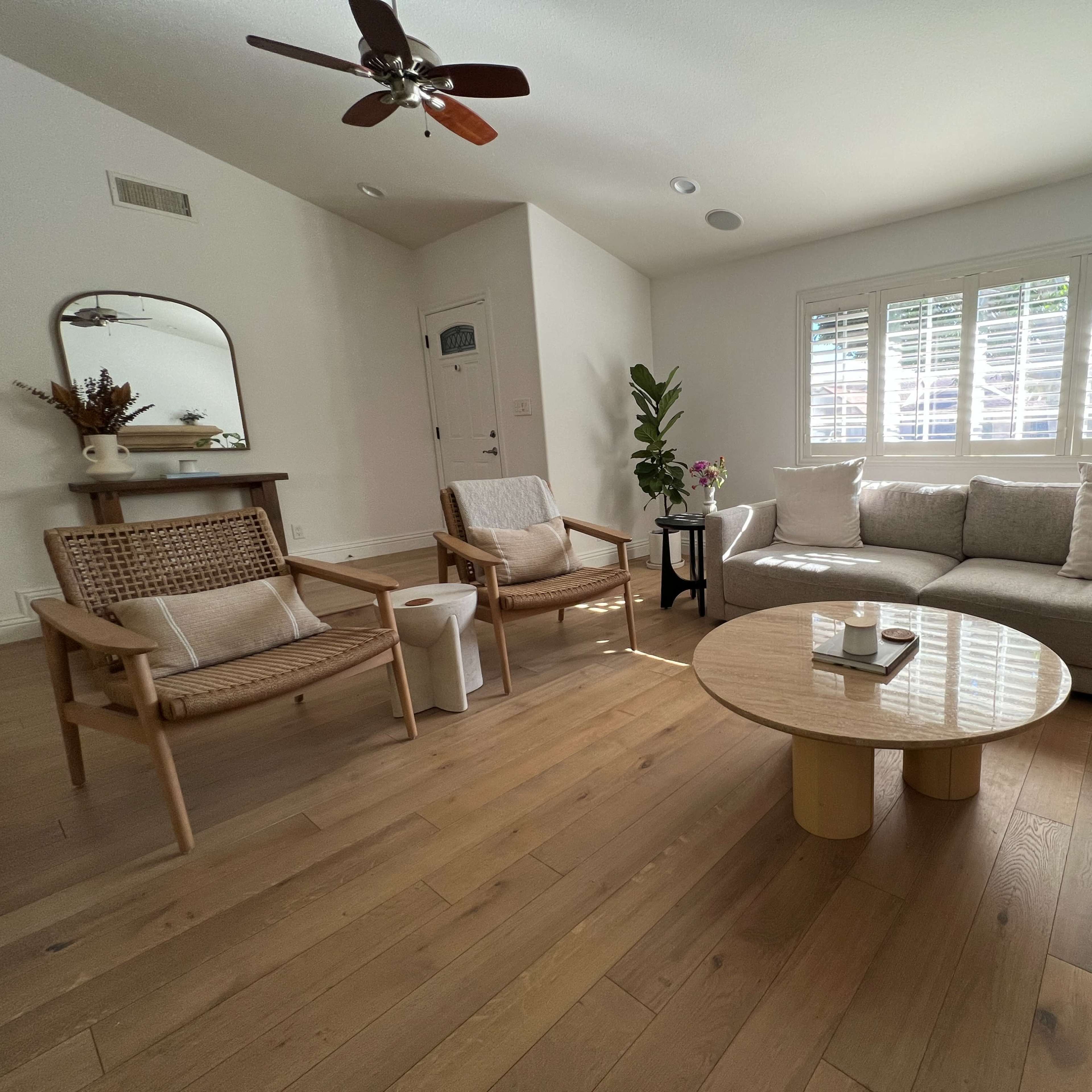 A living room with a light-colored sofa, two woven chairs, a circular coffee table, and bright natural light coming through a window with shutters.