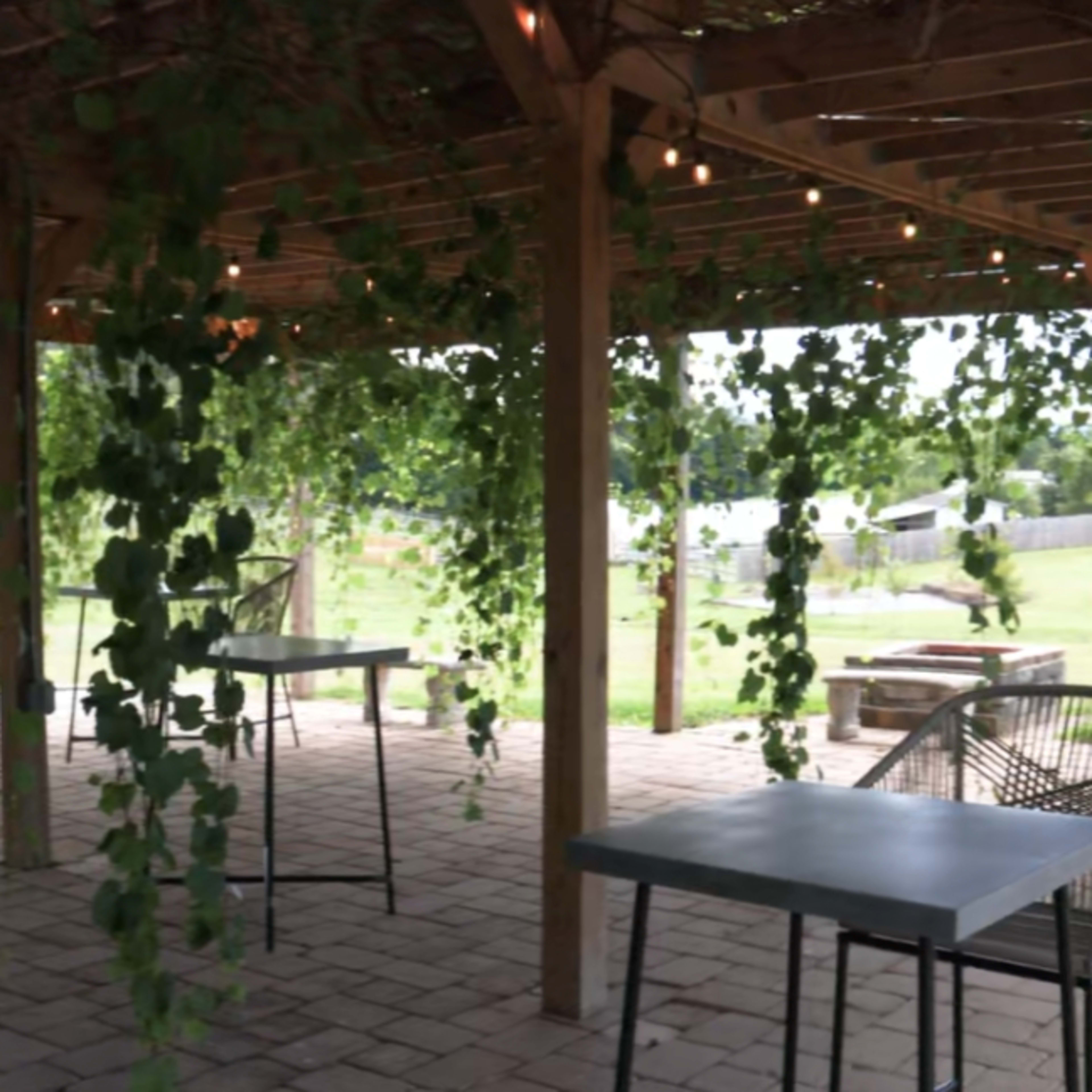 An outdoor dining area with metal tables surrounded by hanging greenery and wooden support beams.