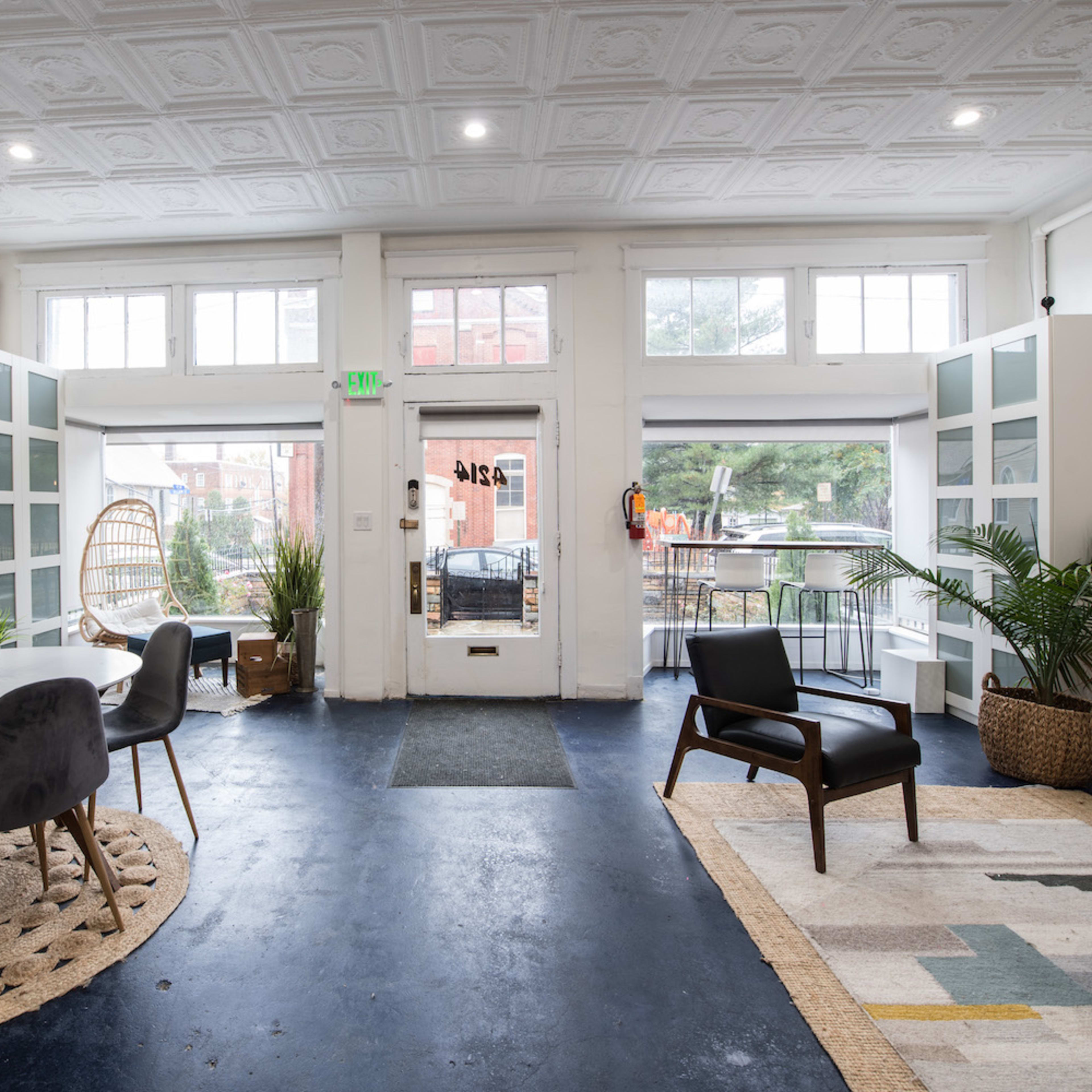 A spacious interior room featuring a circular dining table with chairs, a patterned rug, potted plants, and a seating area with a chair and a sofa.