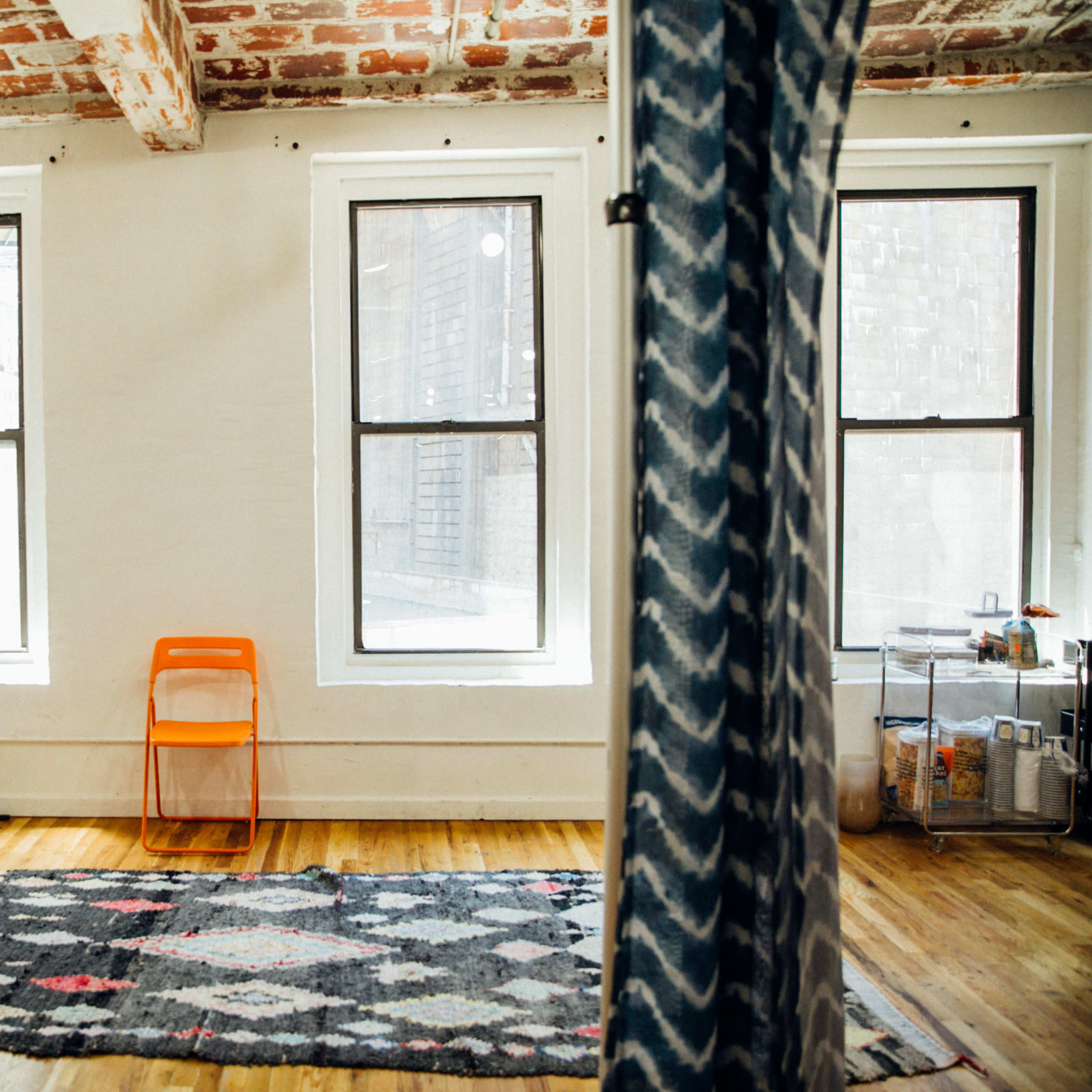 A minimalist room with wooden floors, three windows, an orange chair, a patterned rug, and a mirror against the wall.