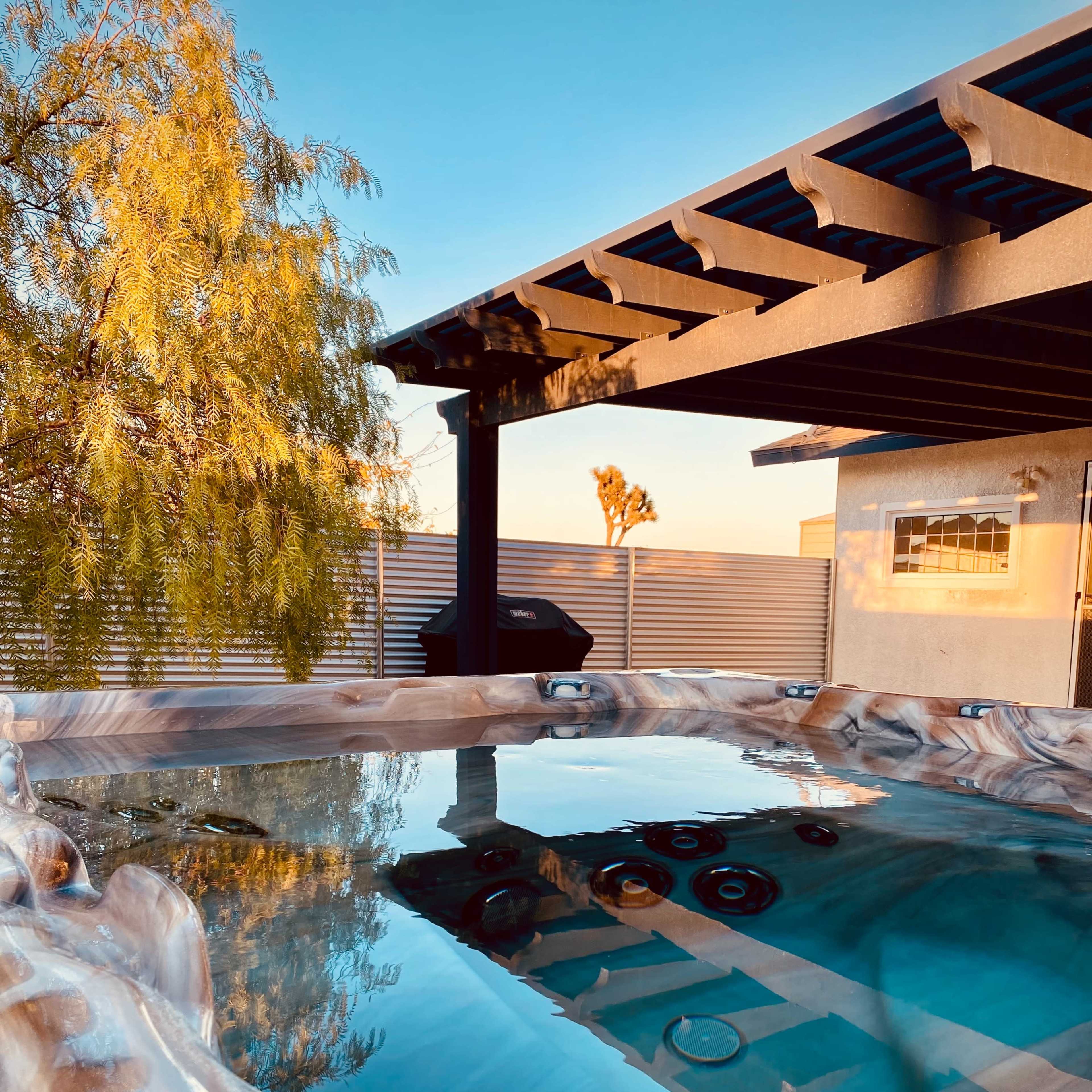 A hot tub sits under a pergola, surrounded by a tree and a clear sky, reflecting the nearby structures.