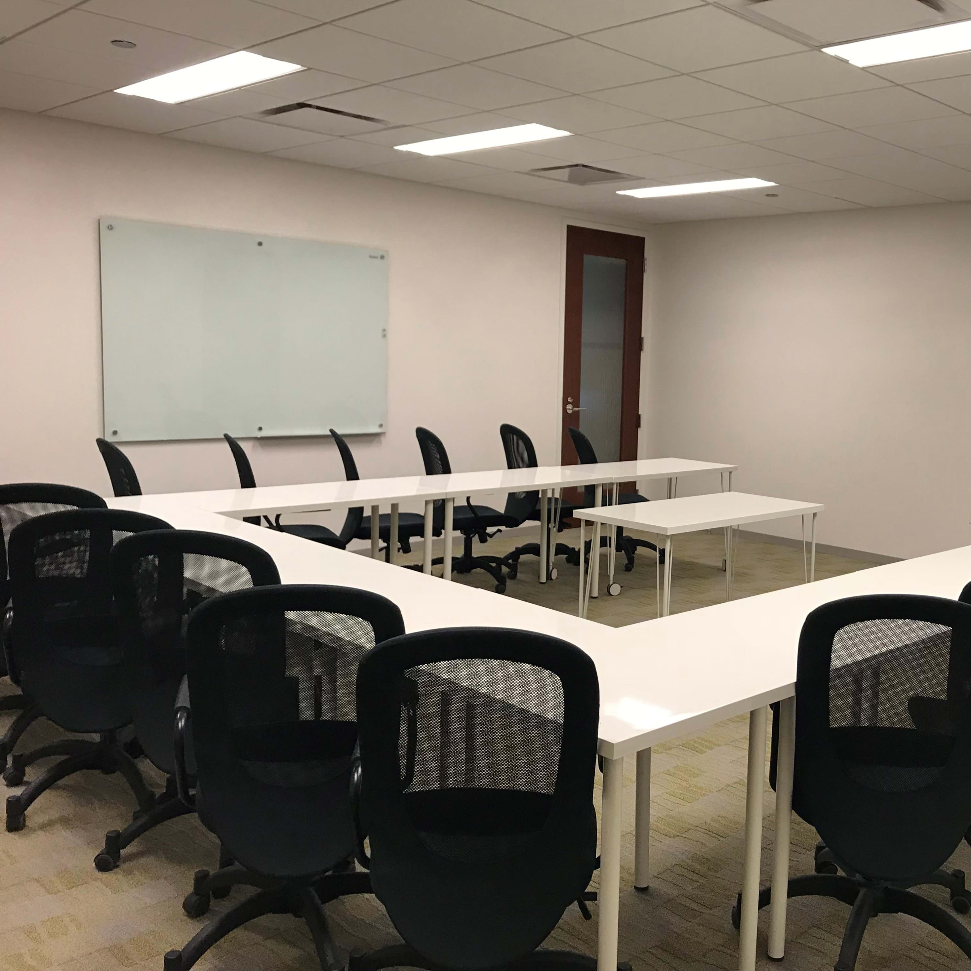 A conference room features a U-shaped arrangement of white tables surrounded by black office chairs, with a whiteboard on one wall.