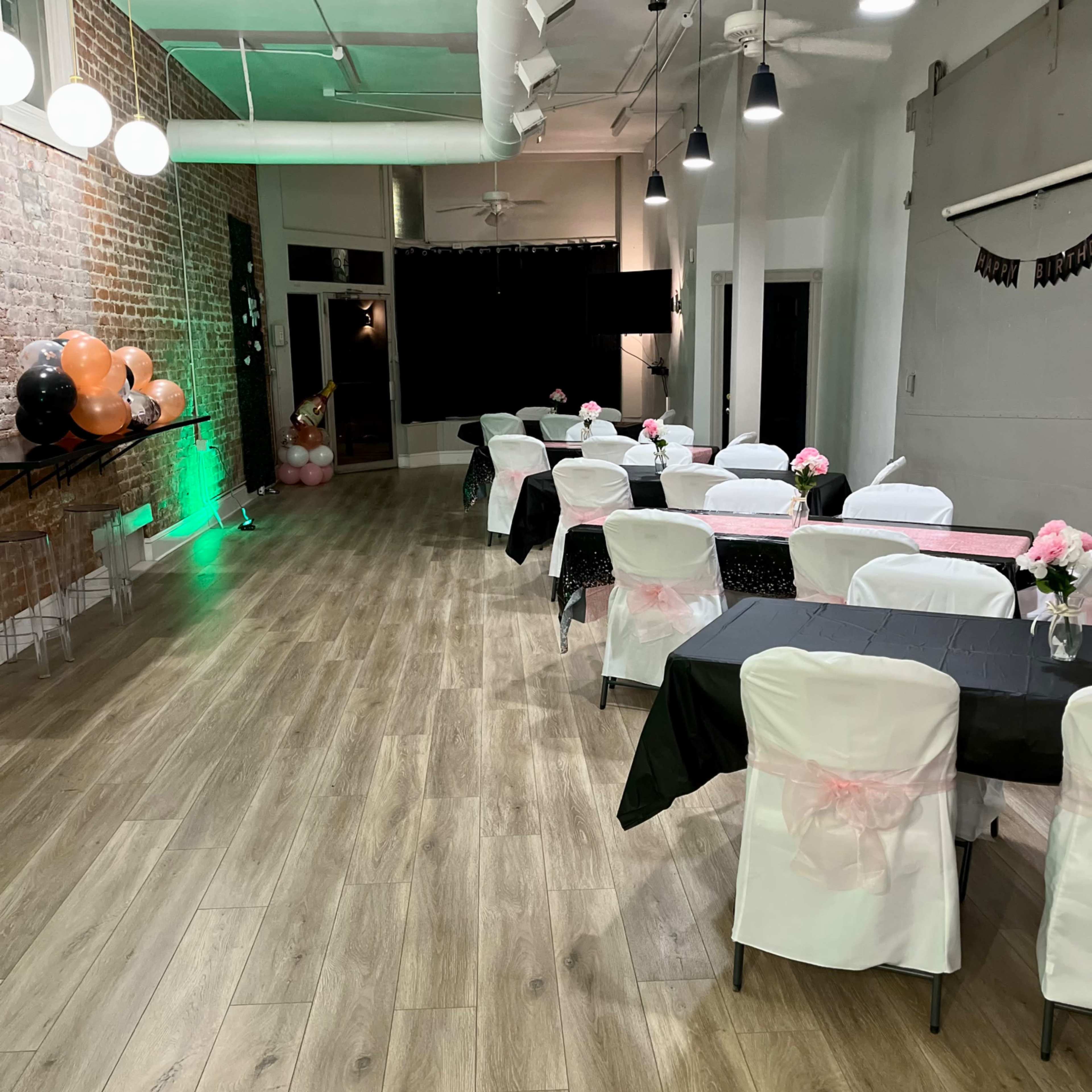A decorated event space with tables set in a row, featuring black and white tablecloths and floral centerpieces, alongside balloons and dim lighting.
