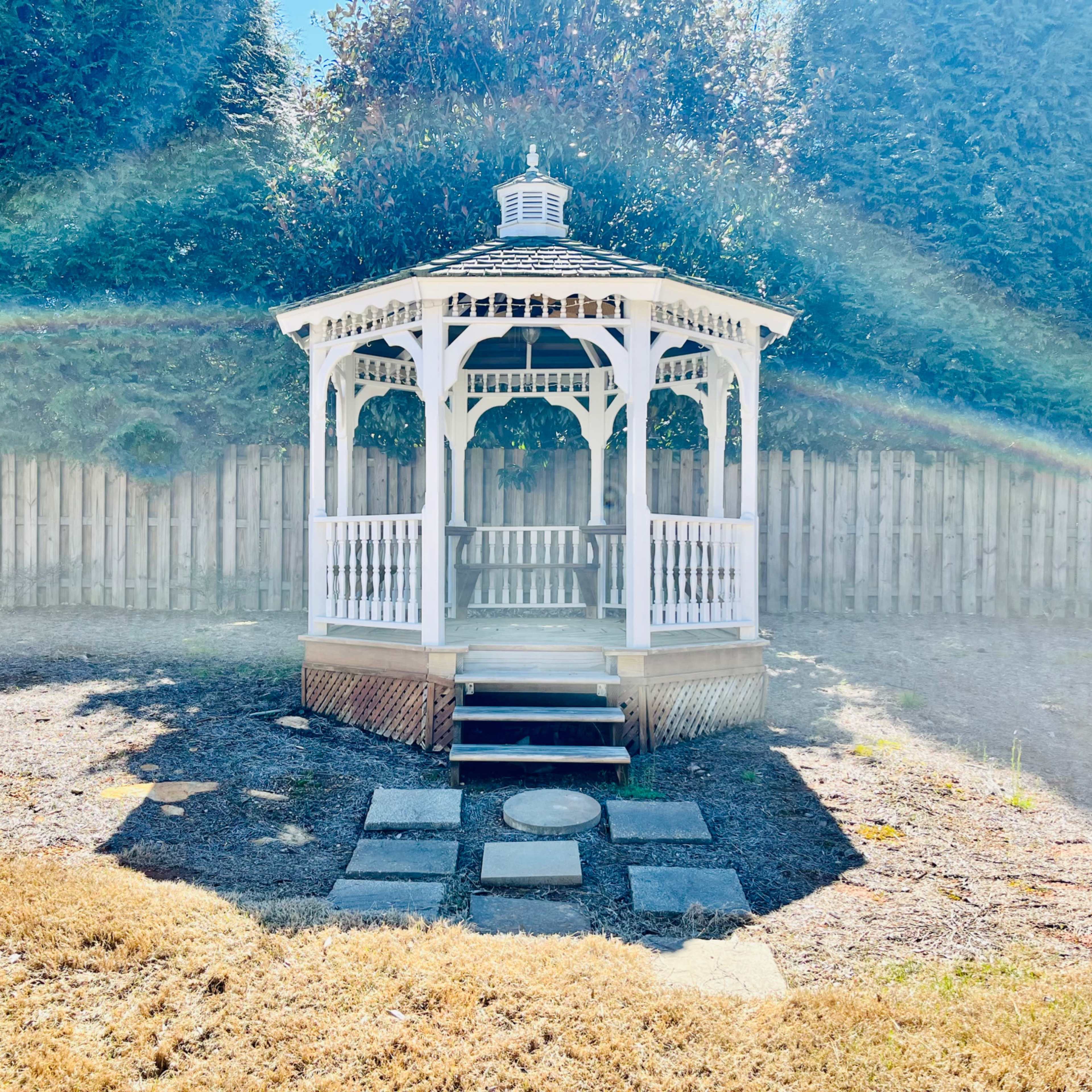 A white gazebo with a shingled roof is positioned on a stone pathway in a grassy yard, surrounded by tall trees and a wooden fence.