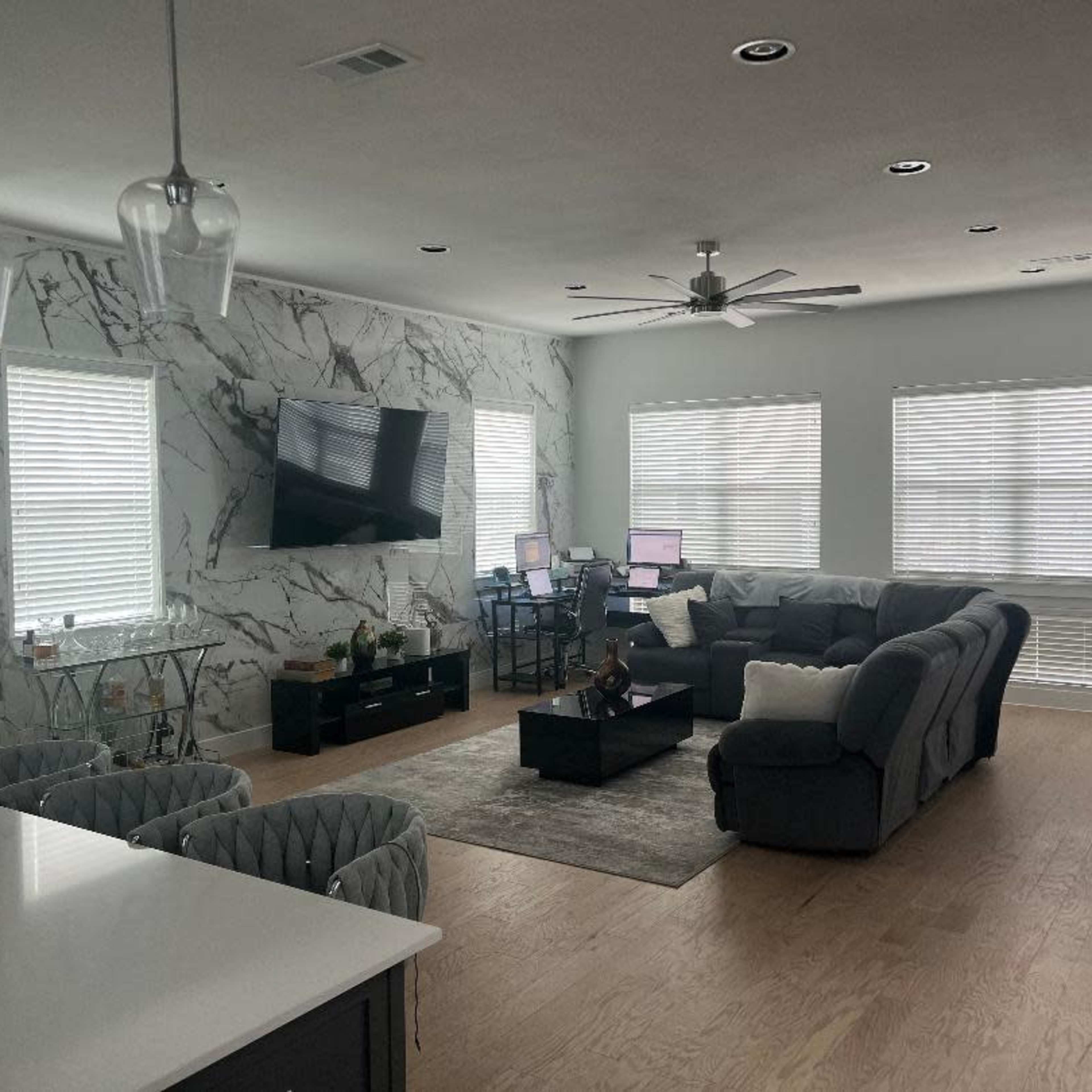 A modern living room with a sectional sofa, a coffee table, a wall-mounted TV, and two workstations against a marble accent wall.