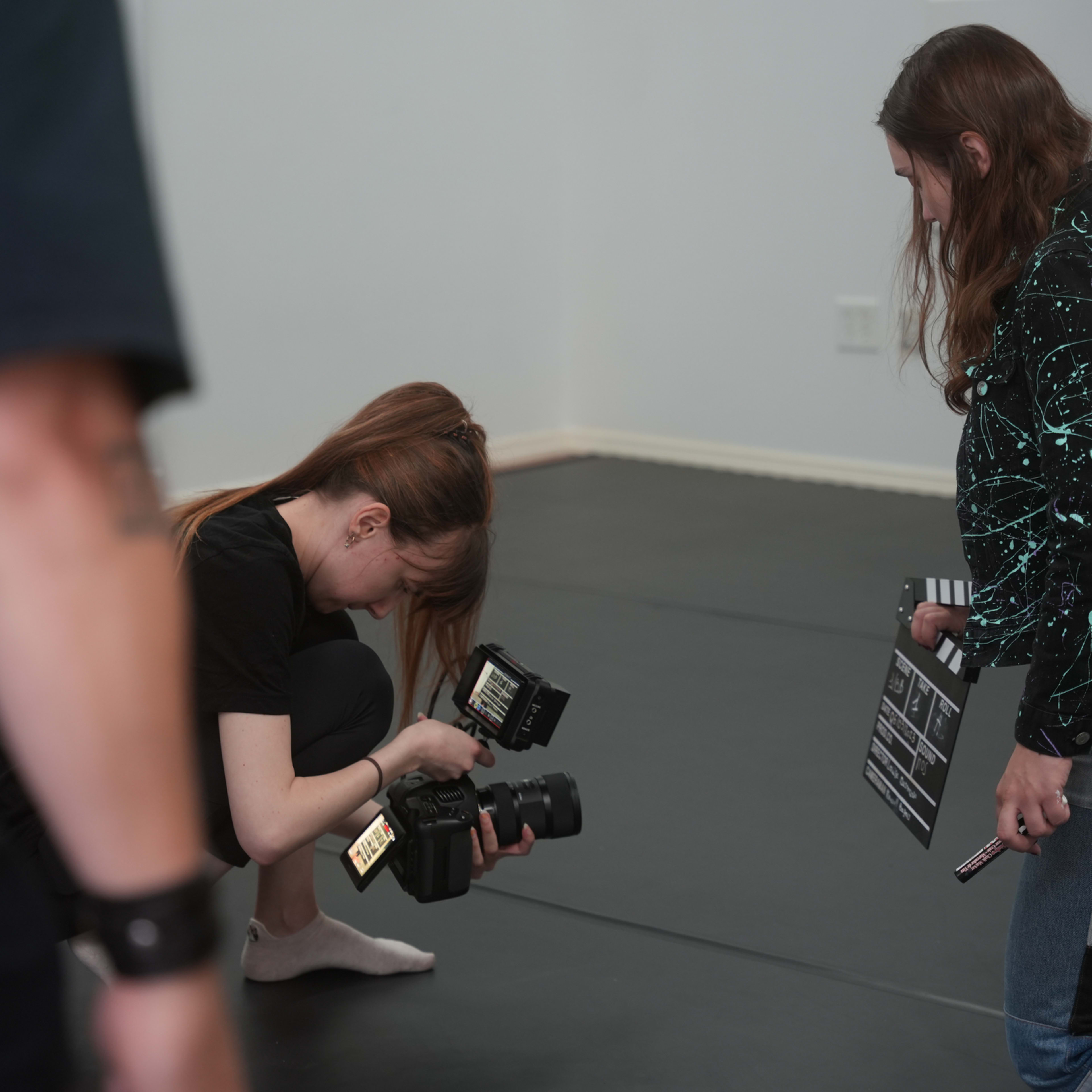 A photographer crouches down to adjust her camera while another person holds a clapperboard on a black mat in a studio setting.