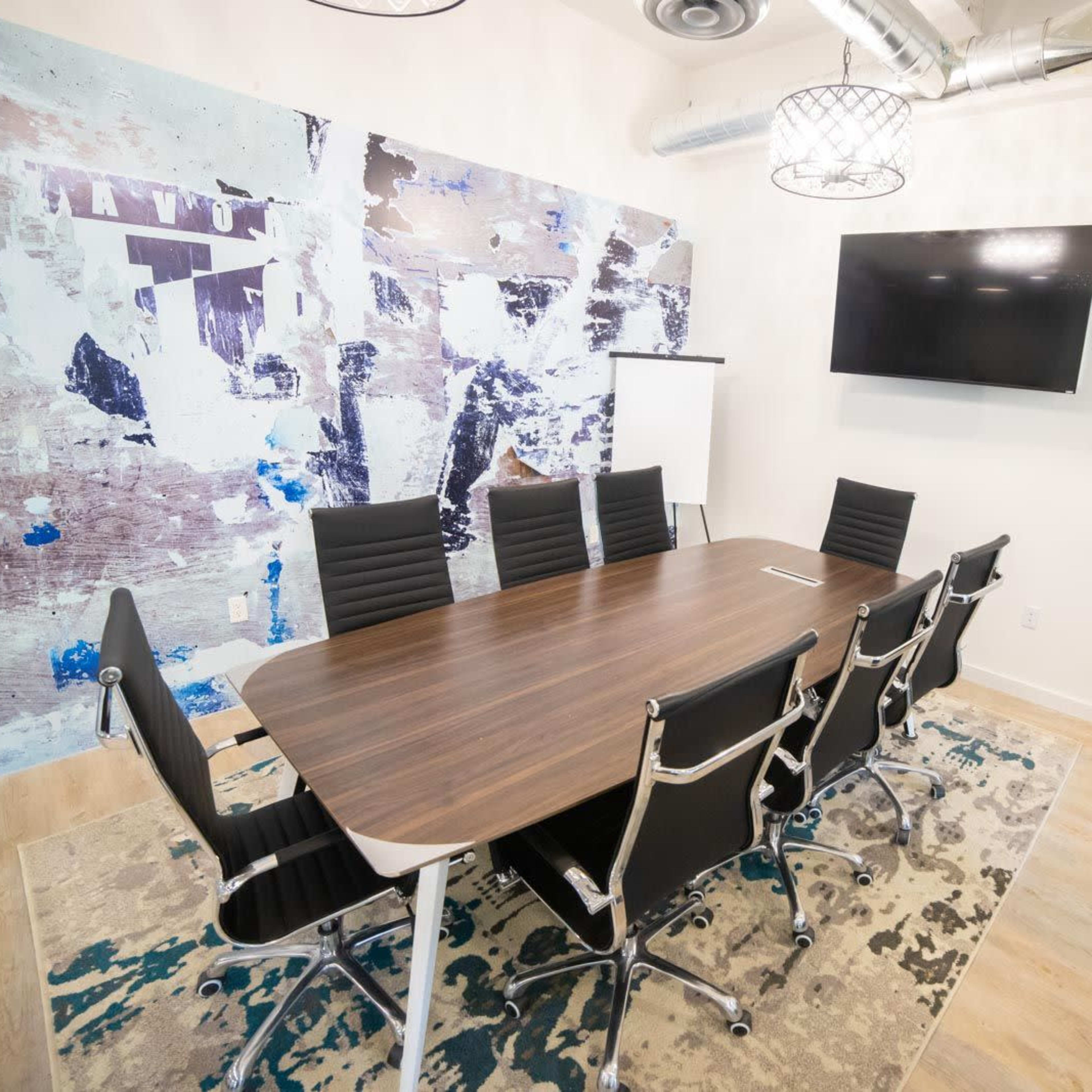 A modern conference room with a large wooden table surrounded by black chairs, a wall featuring abstract artwork, and a television mounted on the wall.