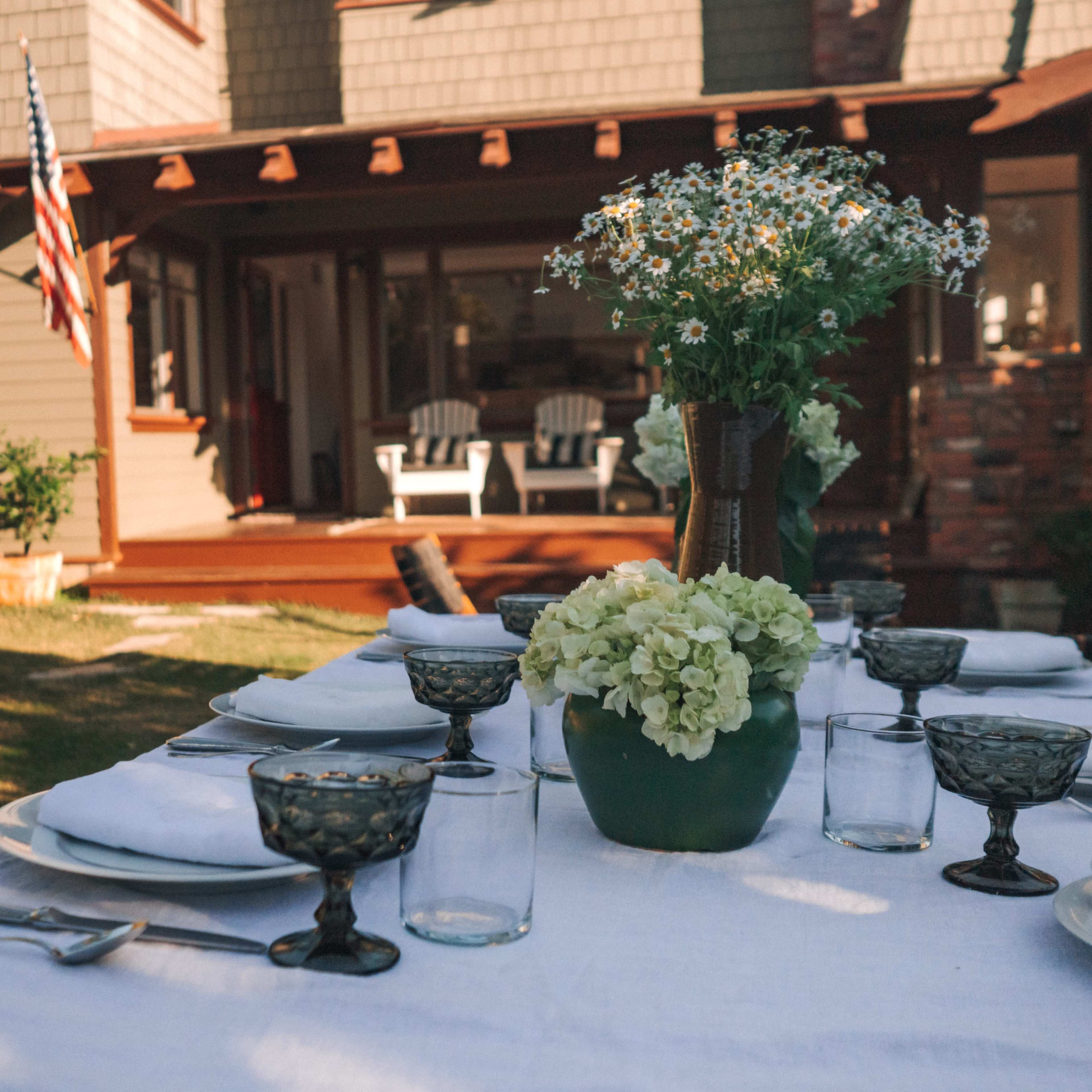 A beautifully set outdoor dining table is arranged with glassware and floral centerpieces in front of a house with a porch.