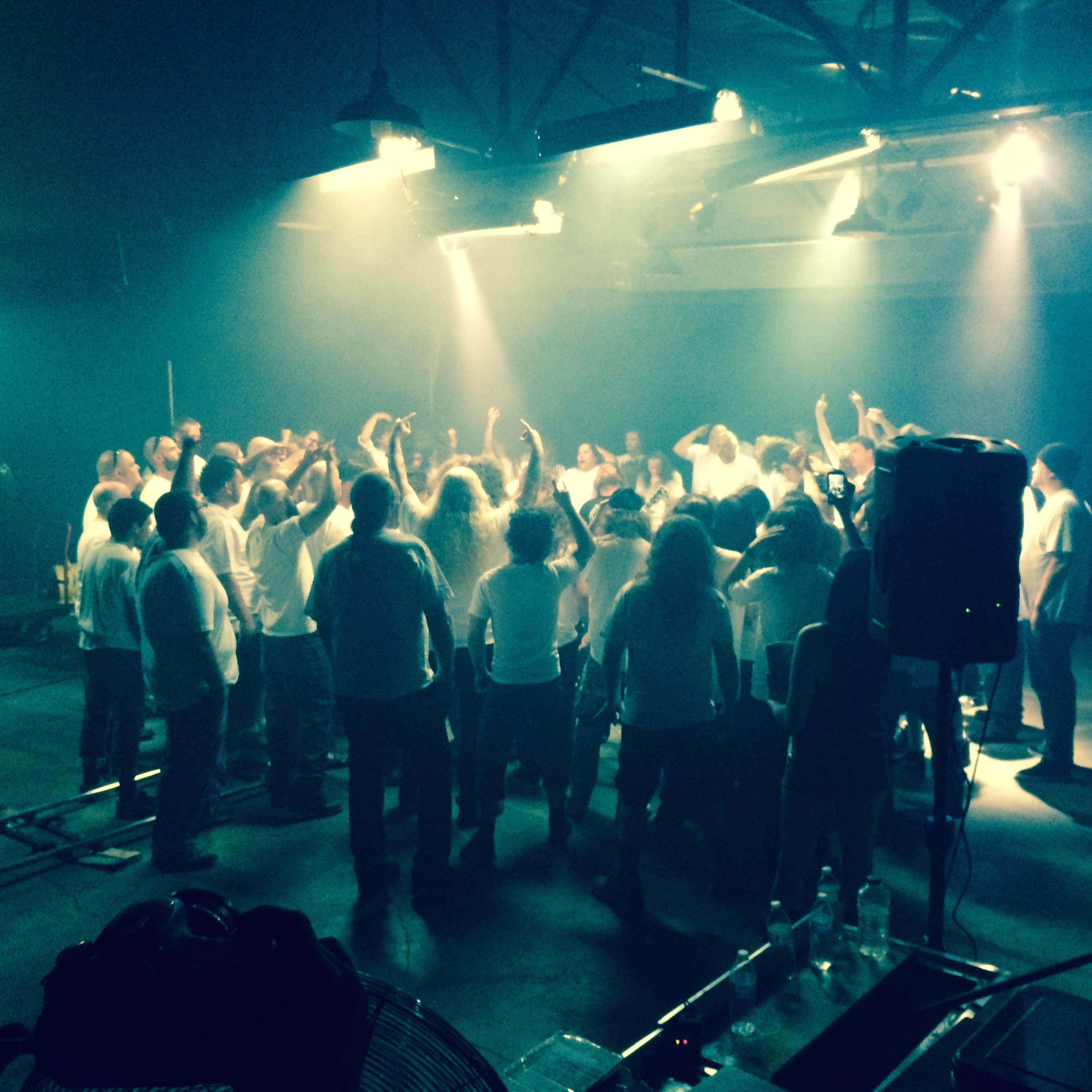 A large group of people in white shirts stands together in a dimly lit space, with raised arms and lights shining down.