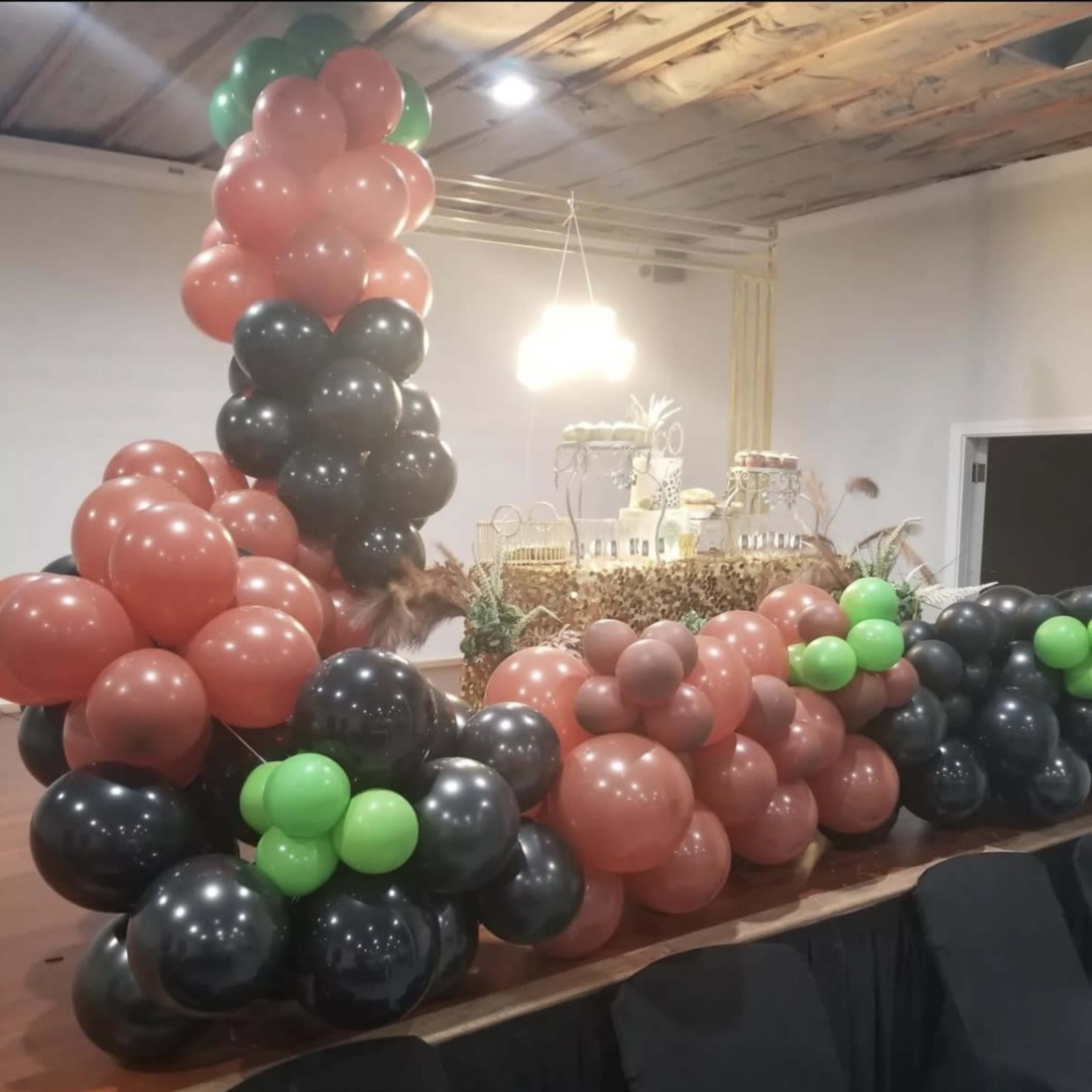 A decorative arrangement of black, green, and brown balloons positioned around a table set with a cake and pastries in a well-lit indoor venue.