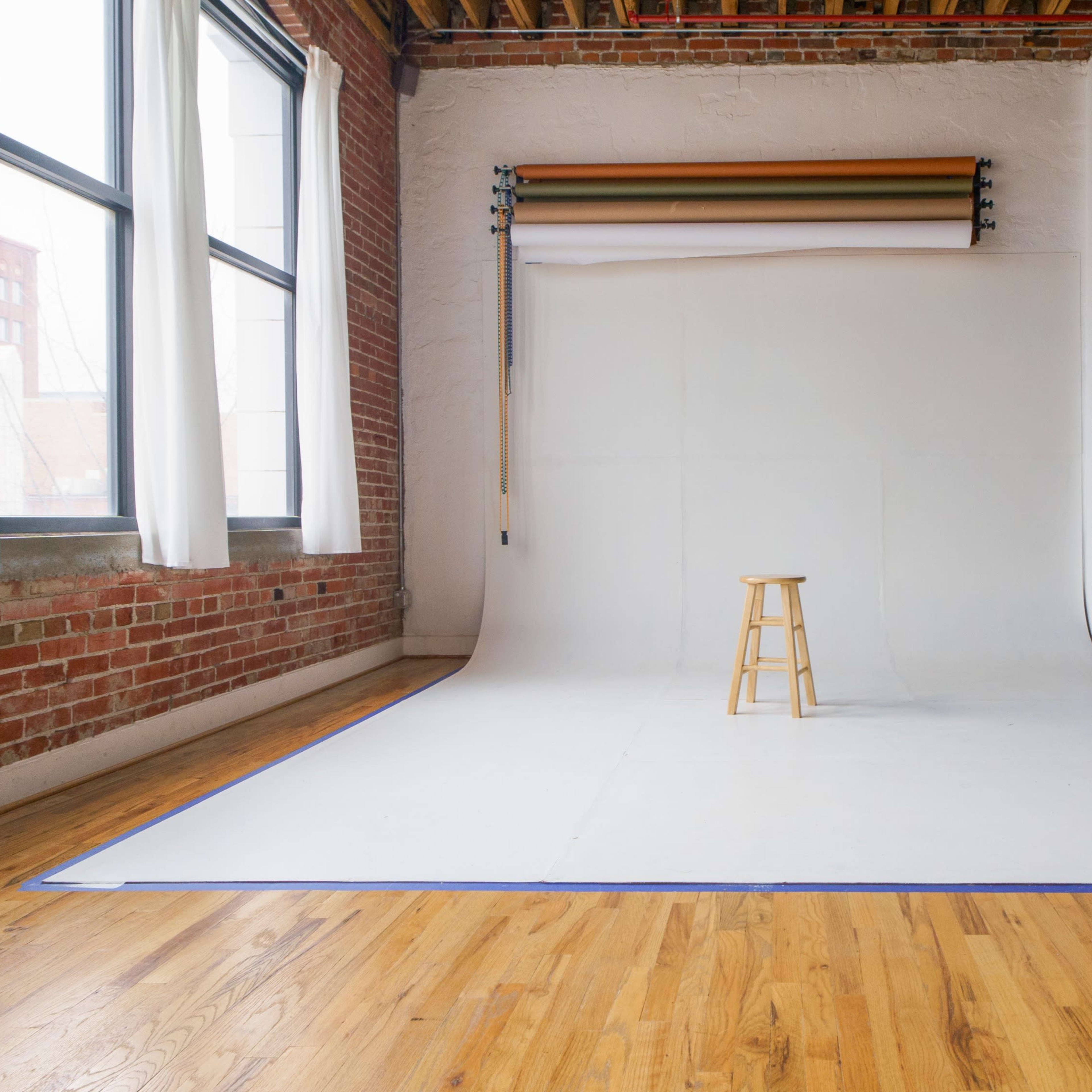 A wooden stool stands on a white backdrop in a bright, spacious room with large windows and exposed brick walls.