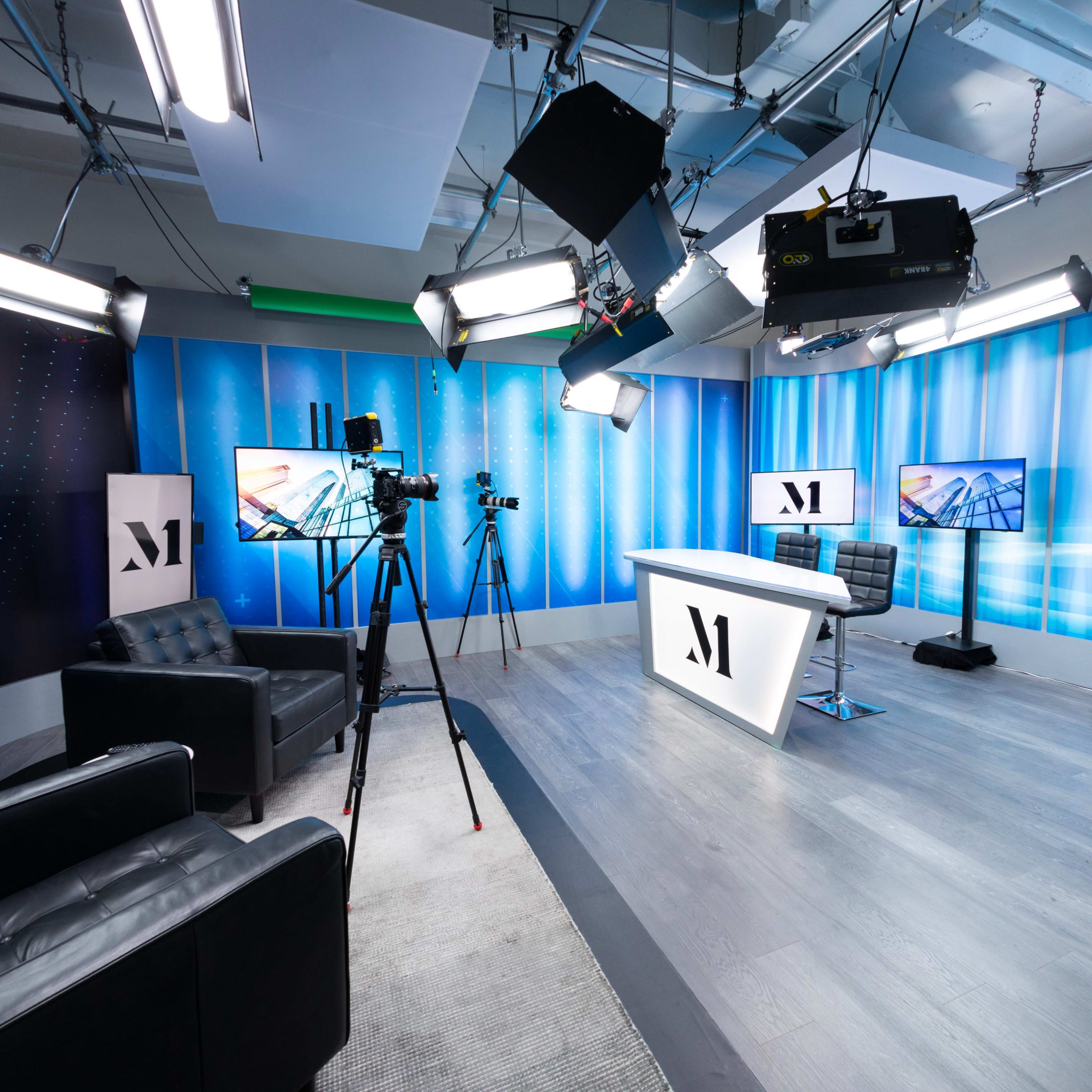 A modern studio setup with multiple cameras, lights, and a central desk surrounded by black leather chairs.