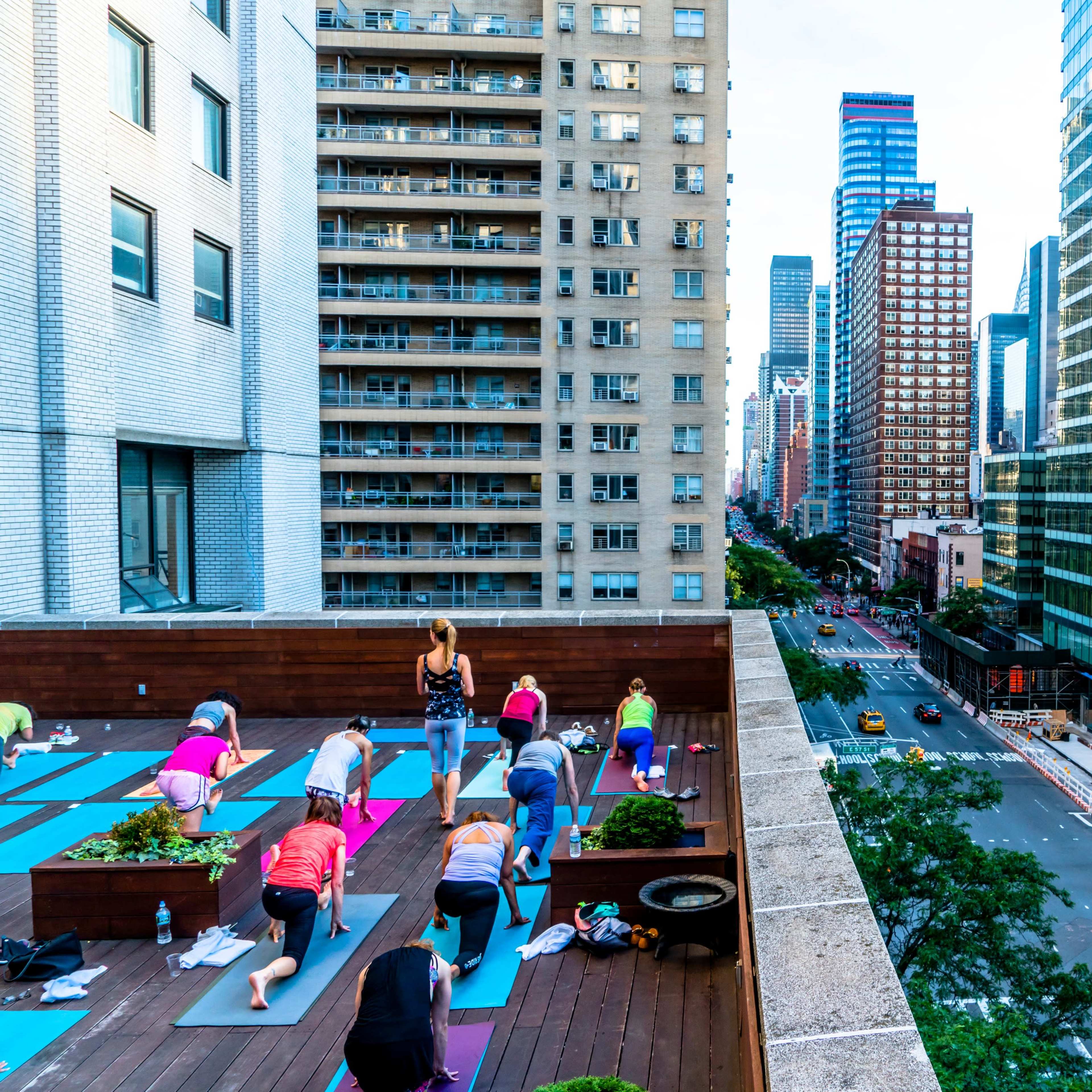 A group of people practices yoga on a rooftop in a city, with tall buildings and a street visible in the background.