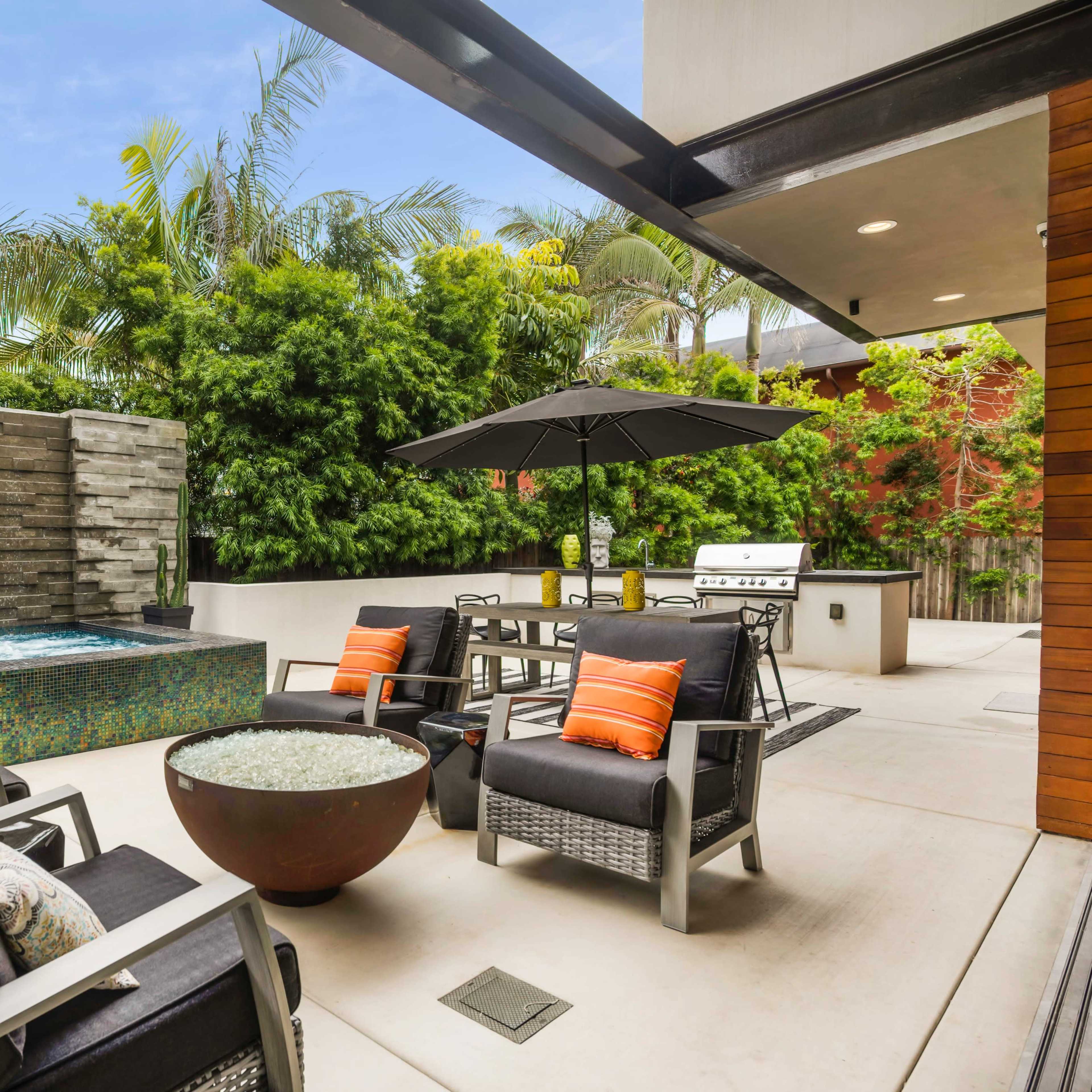A modern outdoor patio area featuring a water fountain, seating with cushions, a dining table under an umbrella, and a built-in grill surrounded by lush greenery.