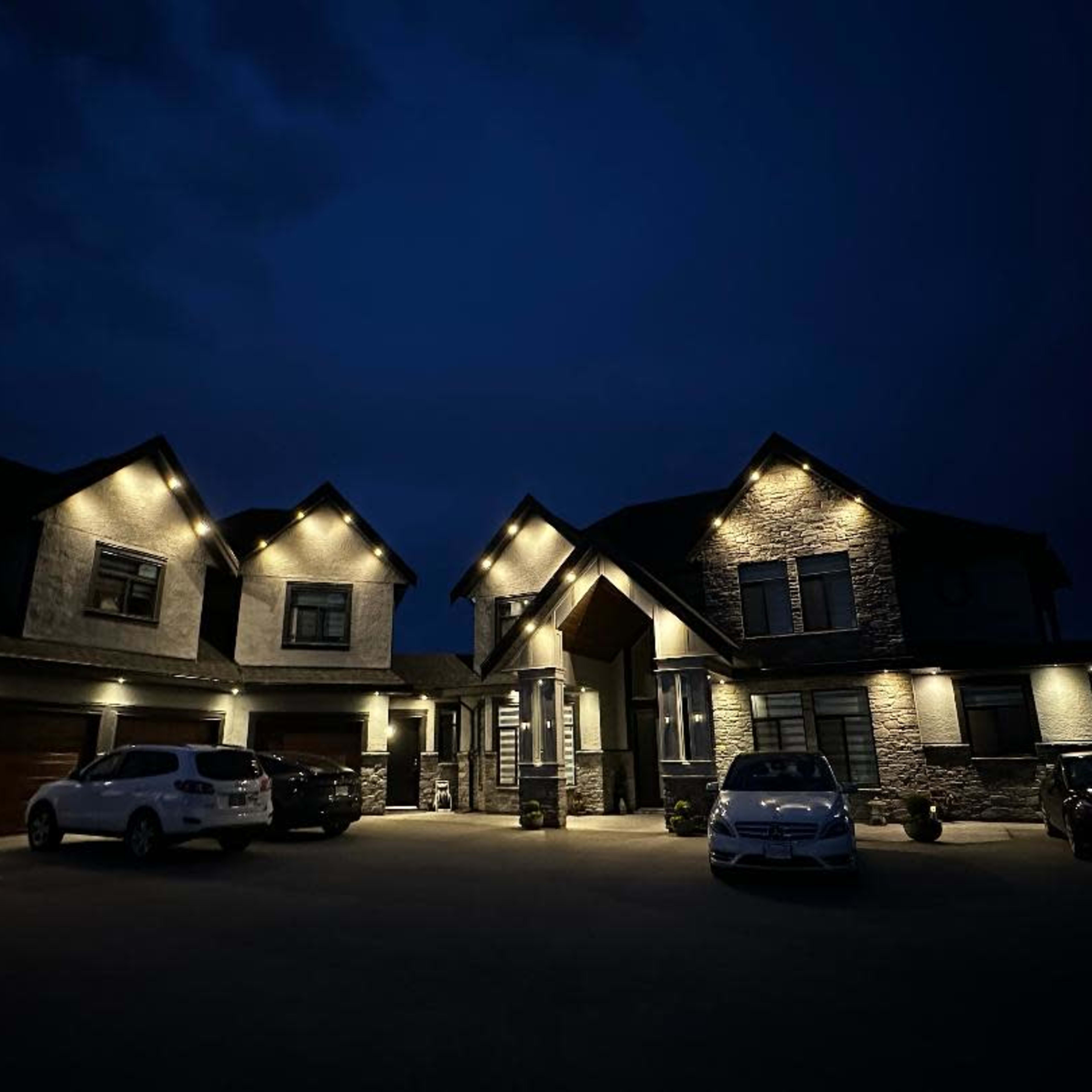 A modern two-story house with stone accents and decorative lighting is situated at night, surrounded by several parked cars in a well-lit driveway.