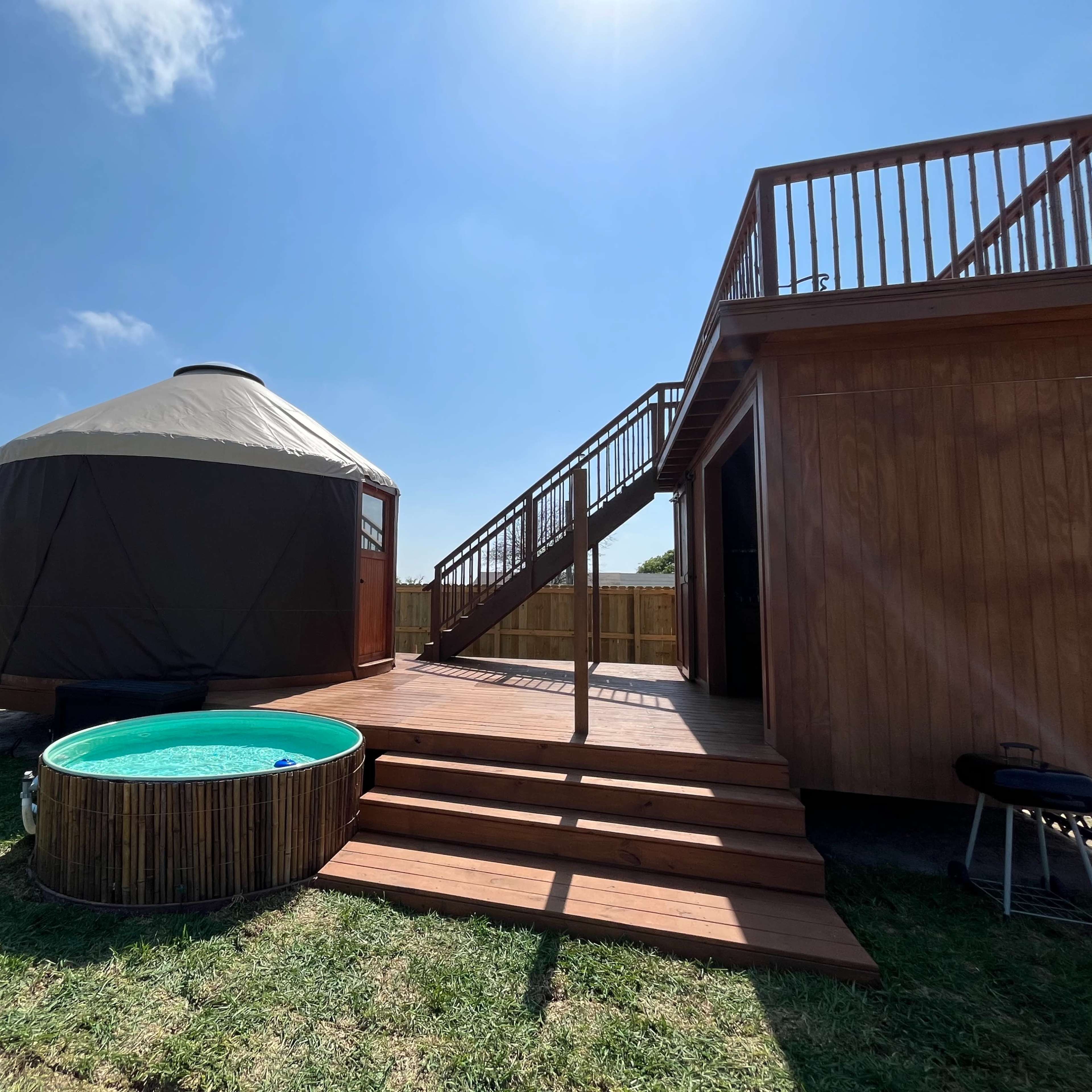 A wooden deck leads to a round yurt and a small above-ground pool in a grassy outdoor setting.