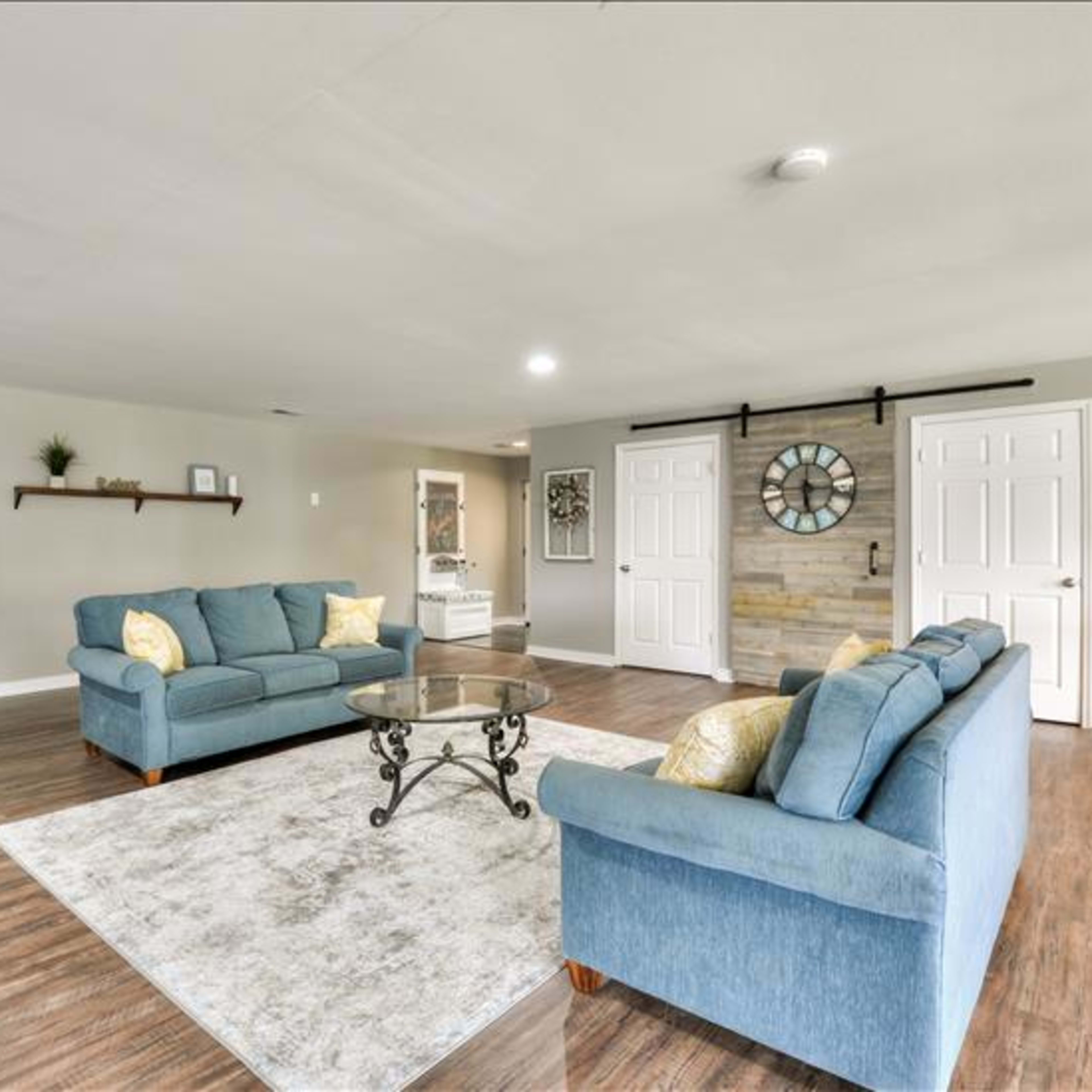 A spacious living room featuring two blue sofas, a glass coffee table, and a decorative wall clock against a wood-paneled accent wall.