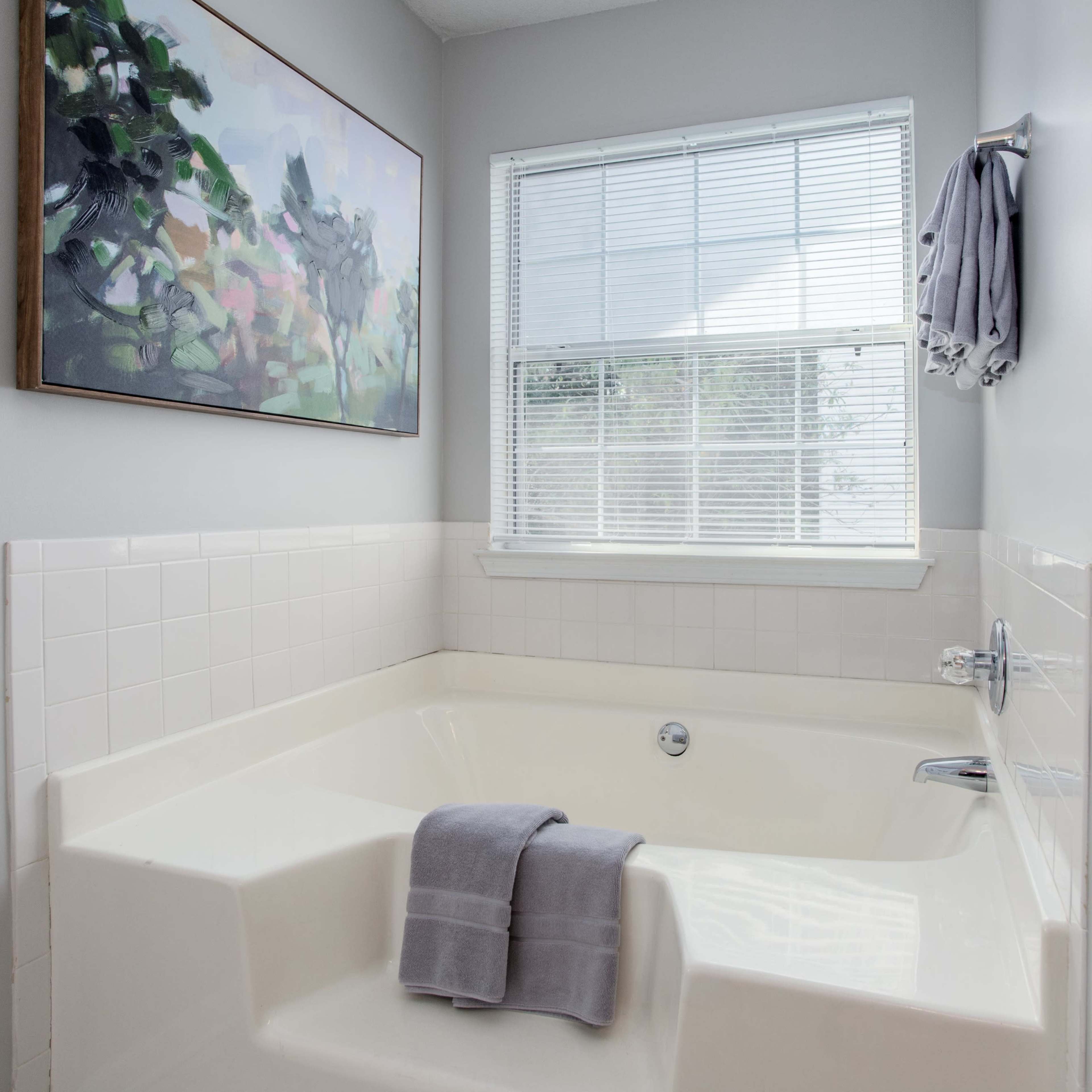 A modern bathroom featuring a corner jetted bathtub, a window, and a shower area, with a piece of artwork and folded towels on display.