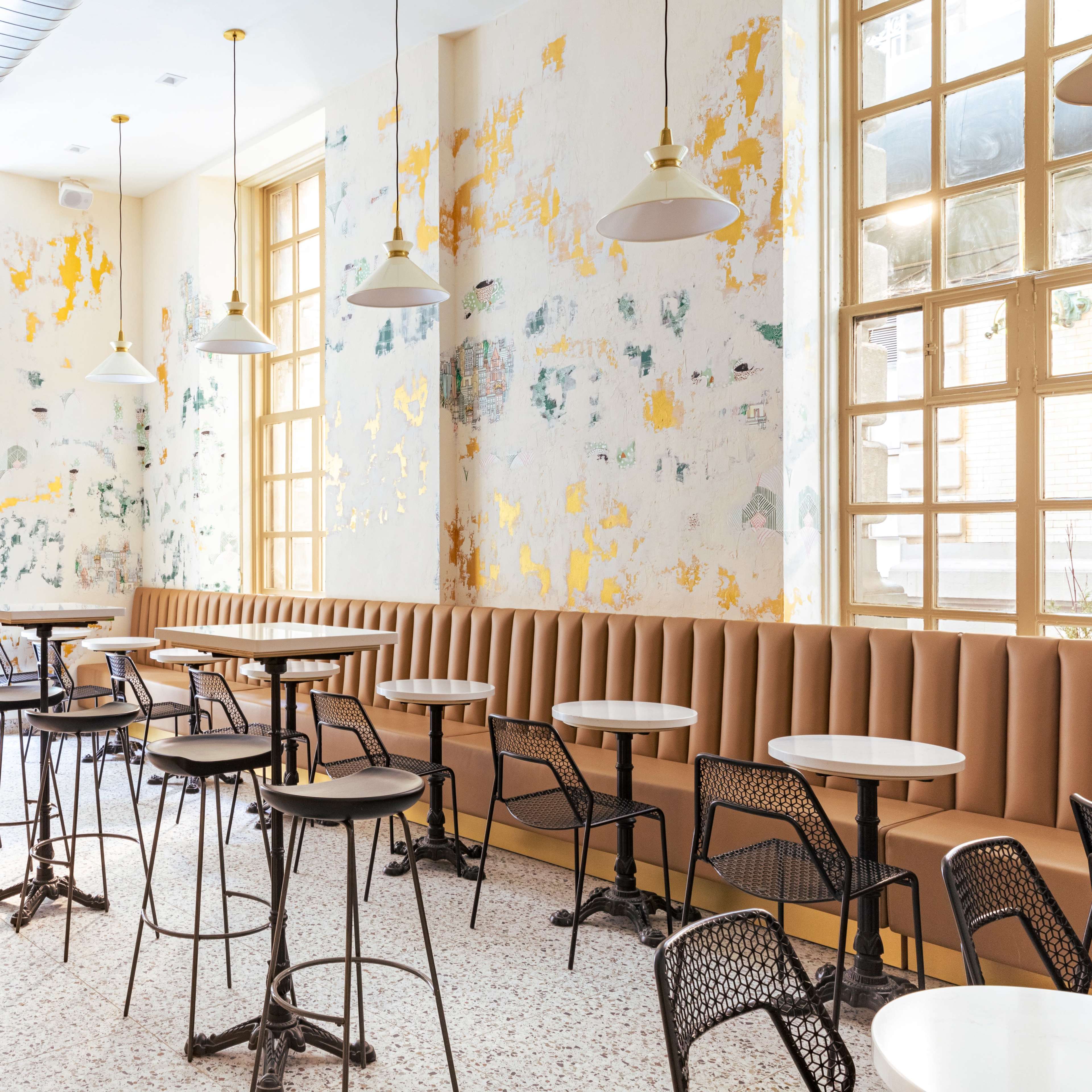 A bright café interior with a mix of brown upholstered seating, round tables, and large windows allowing natural light to enter.