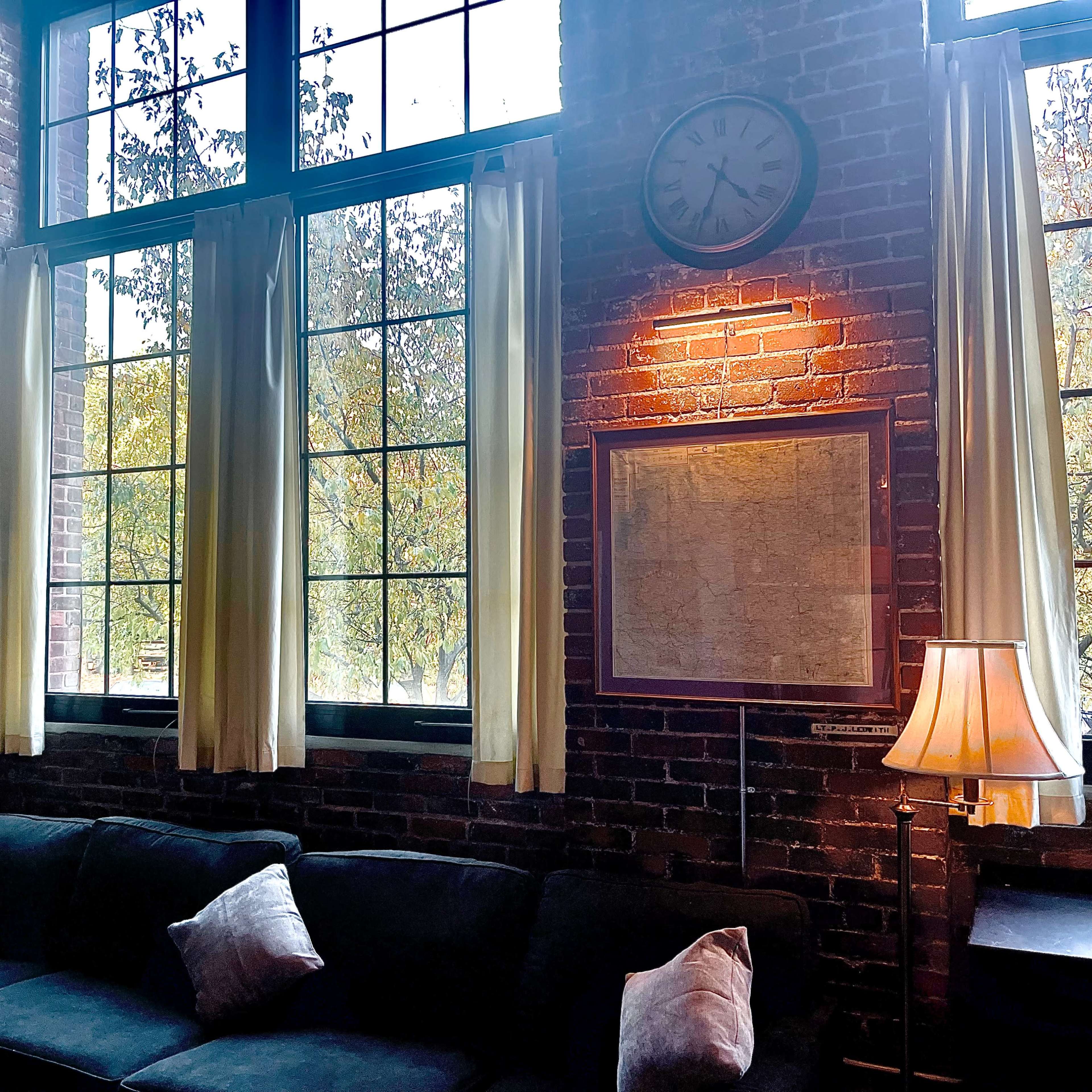 A cozy living area with a dark sofa, large windows framed by light curtains, and a wall clock above a map.