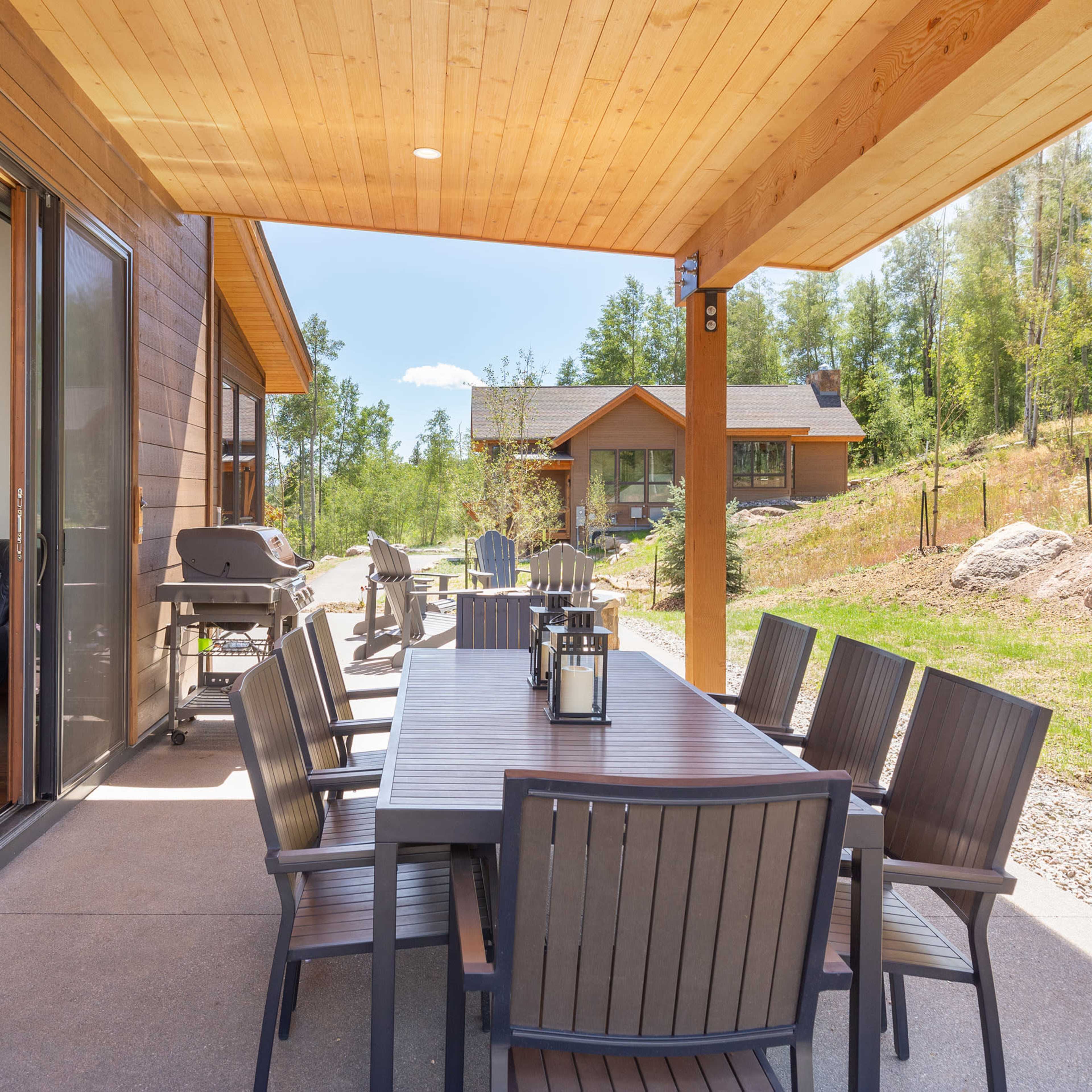A long wooden dining table with chairs sits under a covered patio, overlooking a grassy area and a stone path leading to a house in a wooded setting.