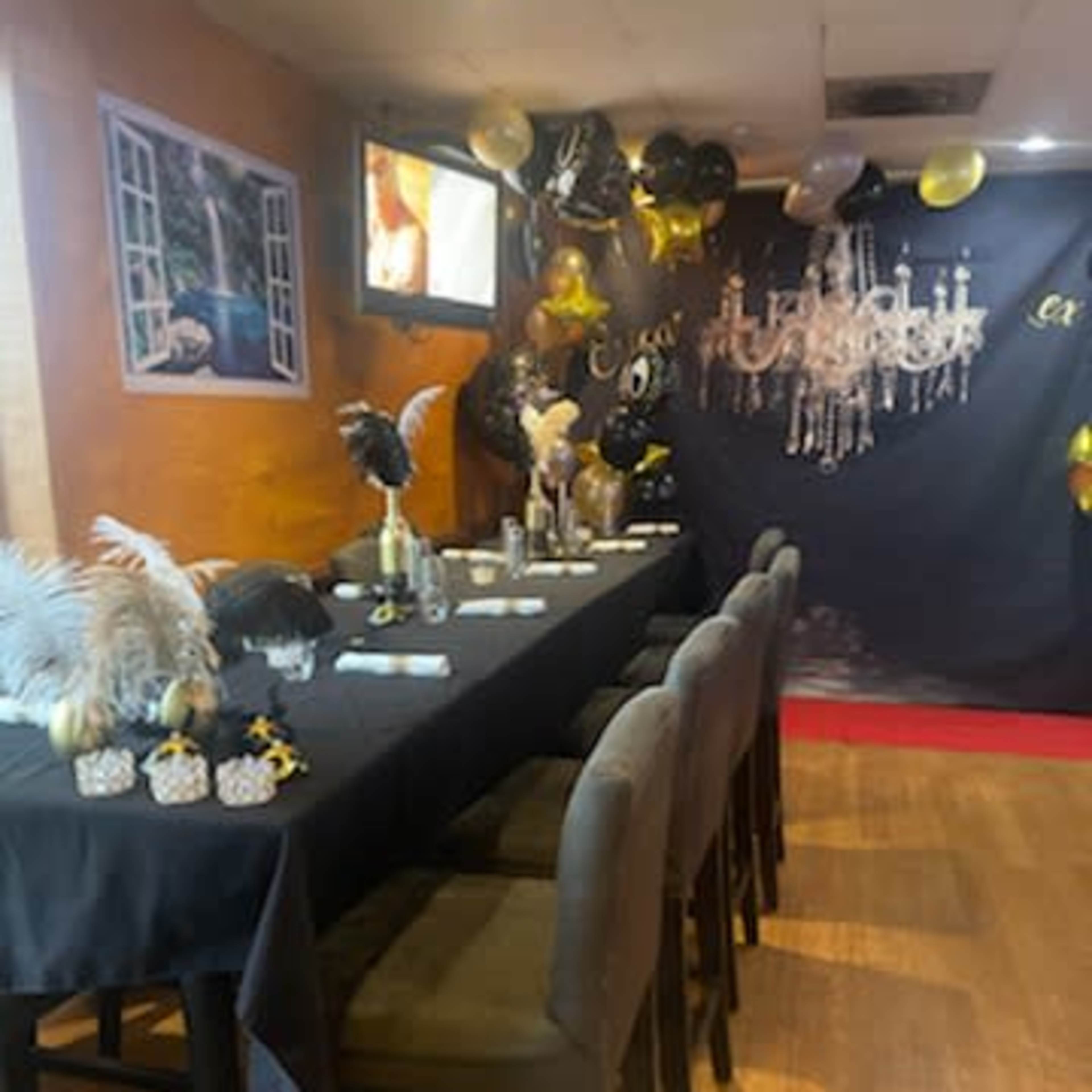 A dining area decorated with black and gold balloons, a long dark table set with utensils, and a background featuring a chandelier and a television screen.