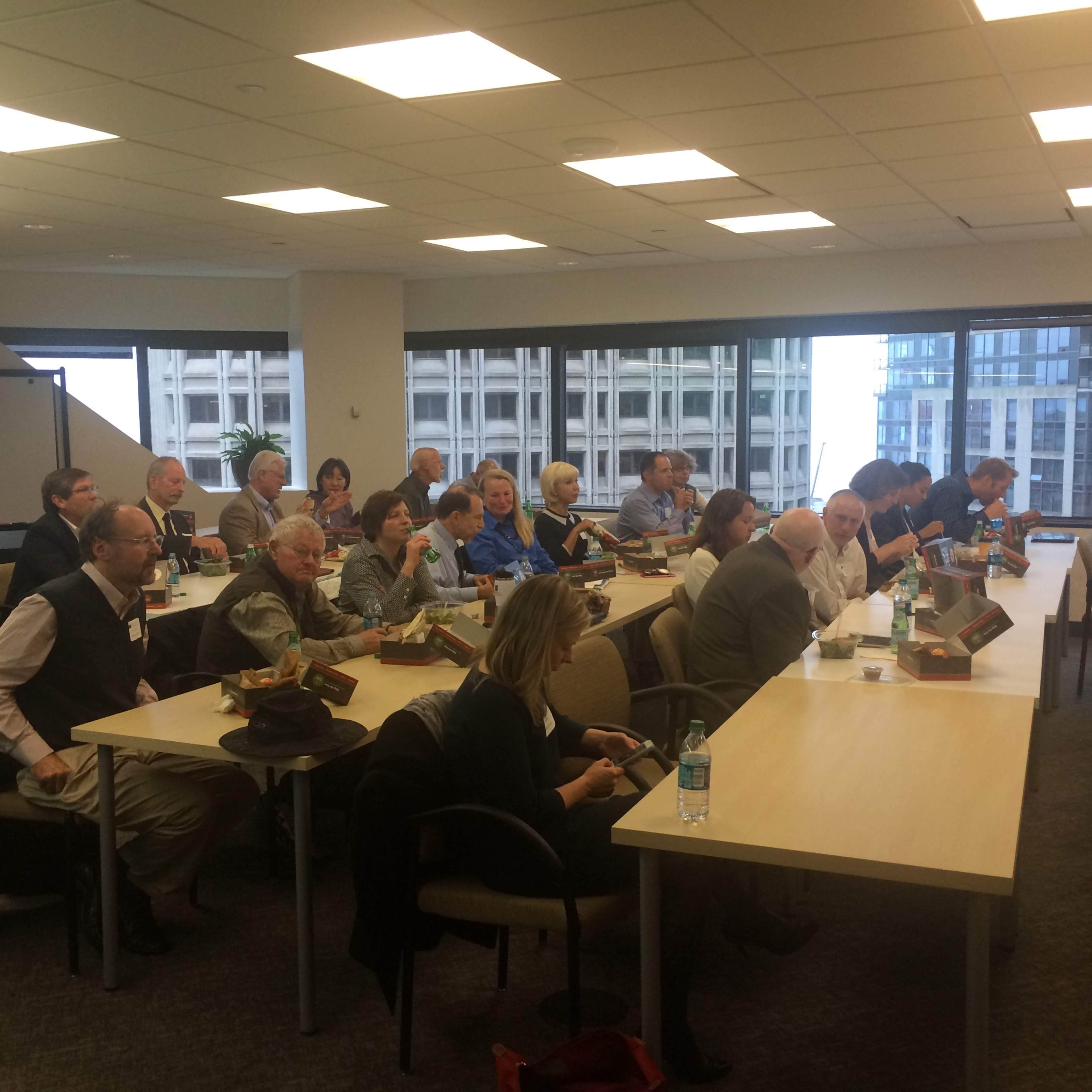 A group of people are seated around tables in a conference room, some looking at electronic devices, while others are engaged in discussion.
