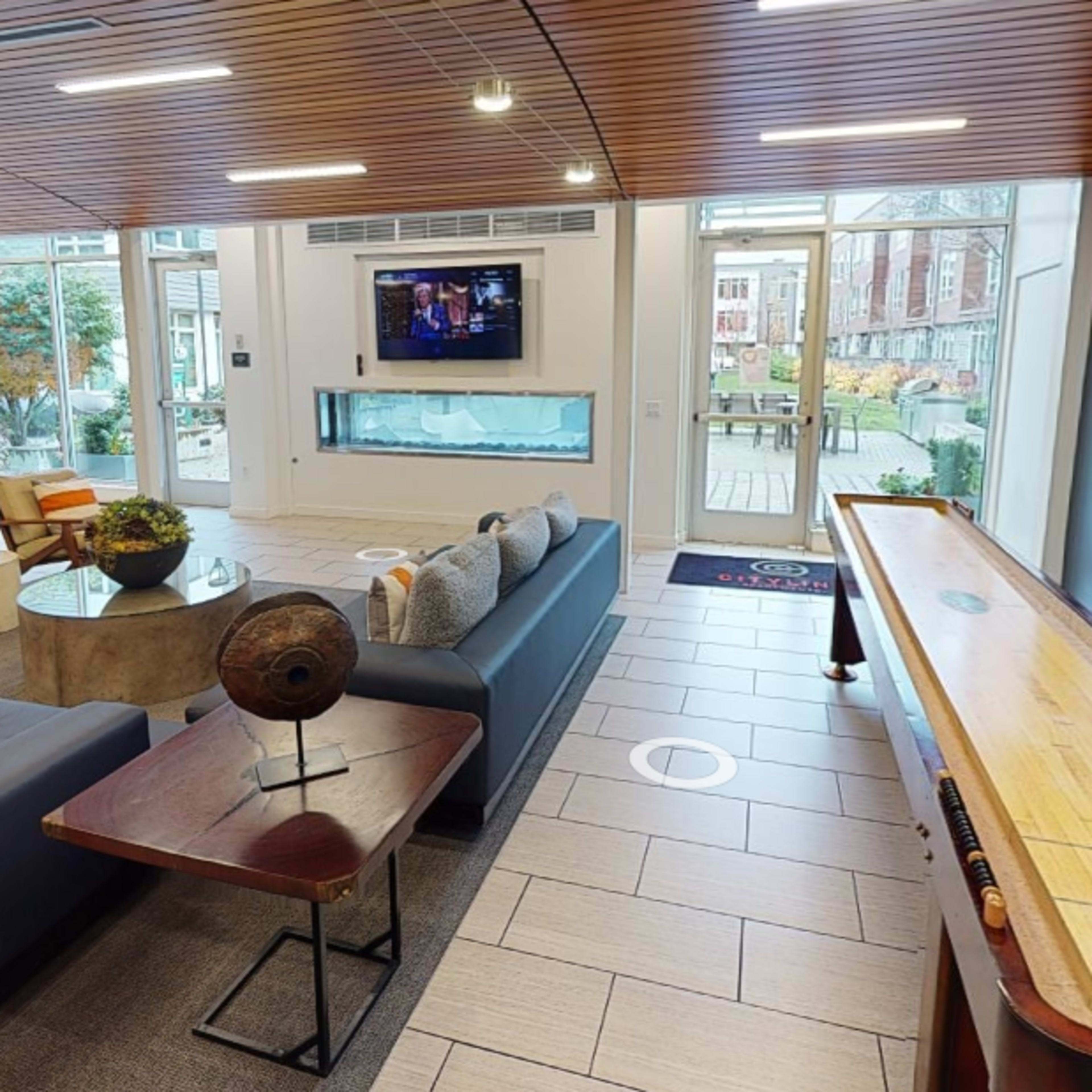 A modern lounge area with a sectional sofa, a coffee table, a television on the wall, and a shuffleboard table, all surrounded by large windows that provide outdoor views.