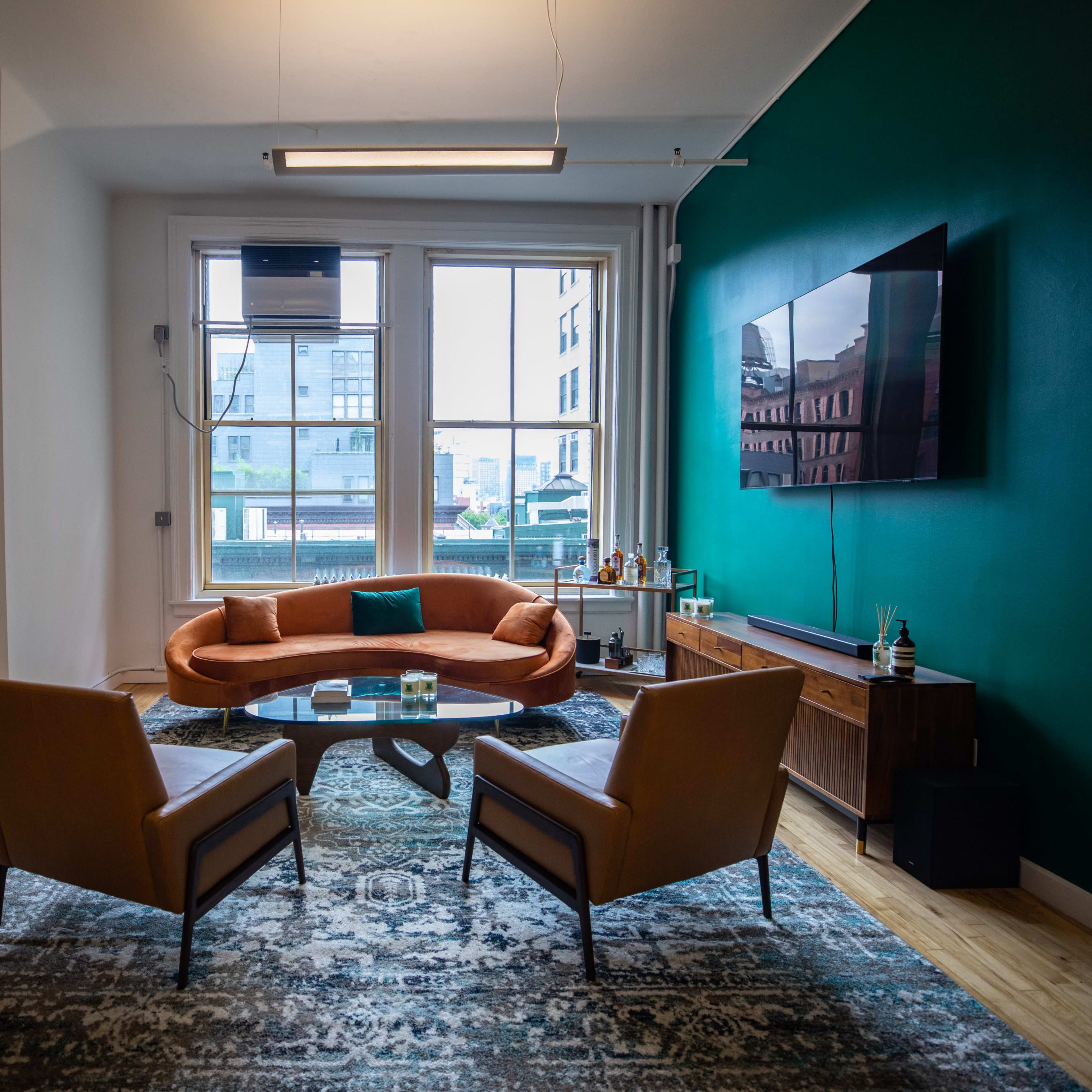 A modern living room with a curved orange sofa, two brown chairs, a coffee table, and a wall-mounted television, all set against a green accent wall and large windows.
