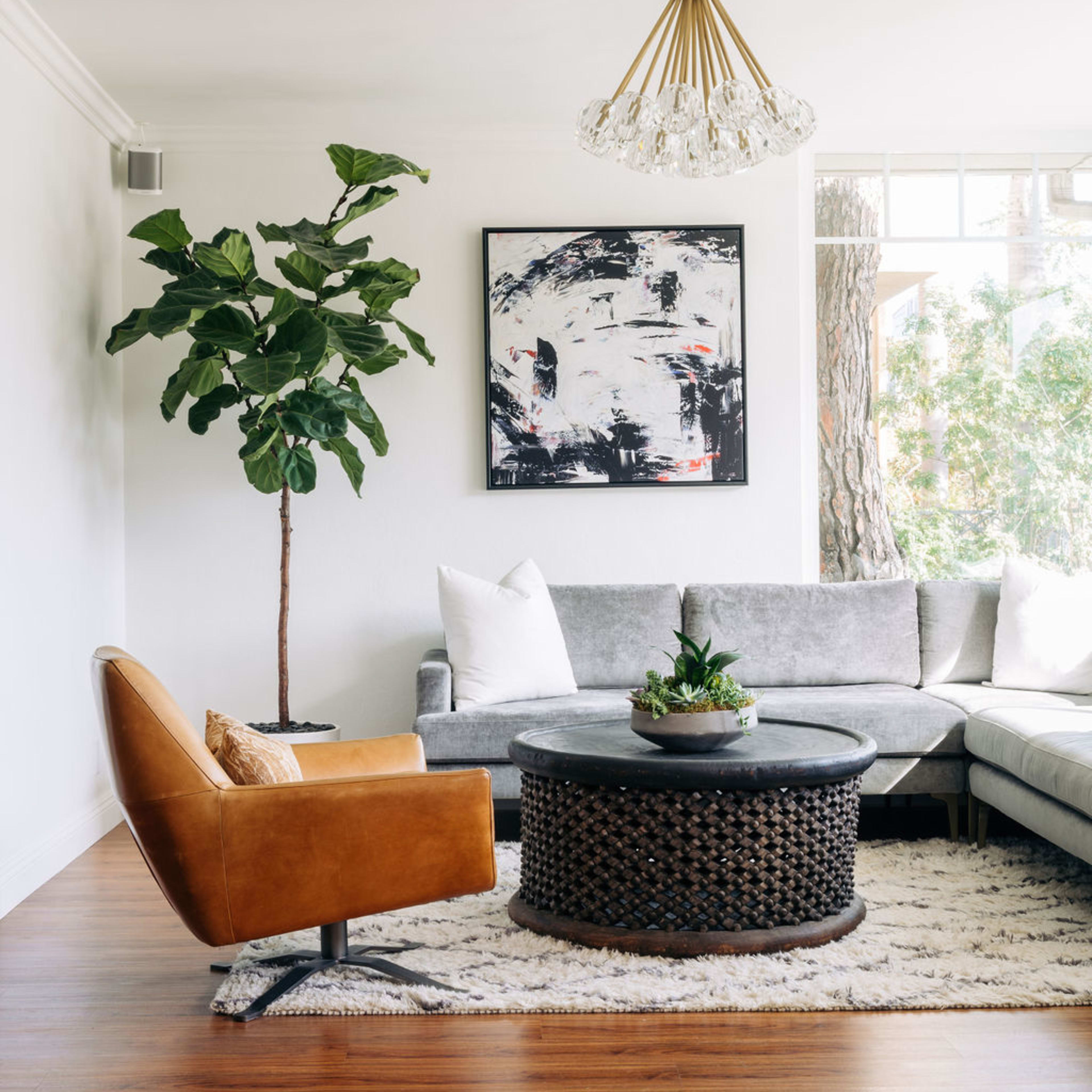 A modern living room features a gray sectional sofa, a brown accent chair, a round coffee table with a woven design, and a tall potted plant, all under a chandelier.