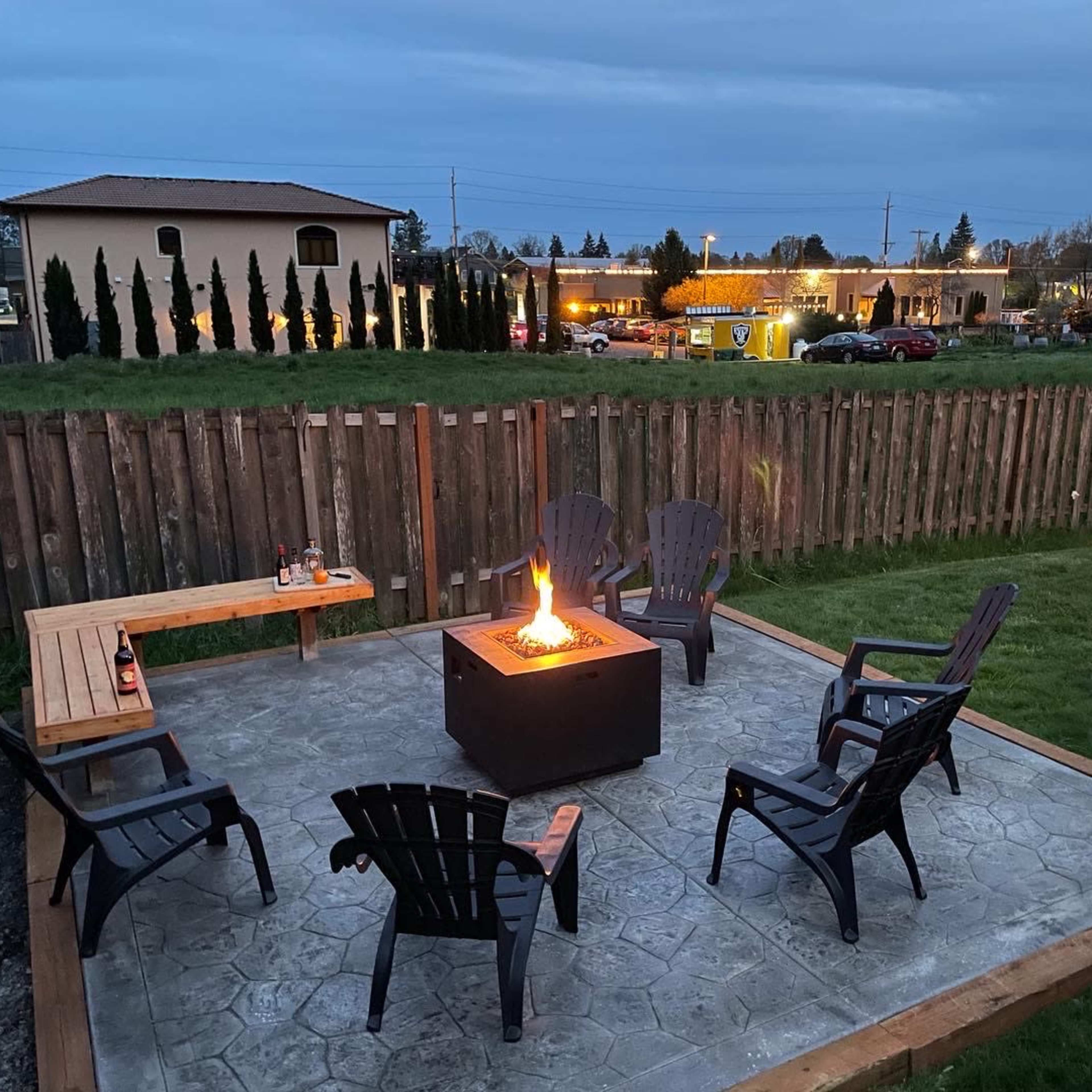 A firepit surrounded by six outdoor chairs is situated on a concrete patio, with a wooden bench and a wooden fence in the background.