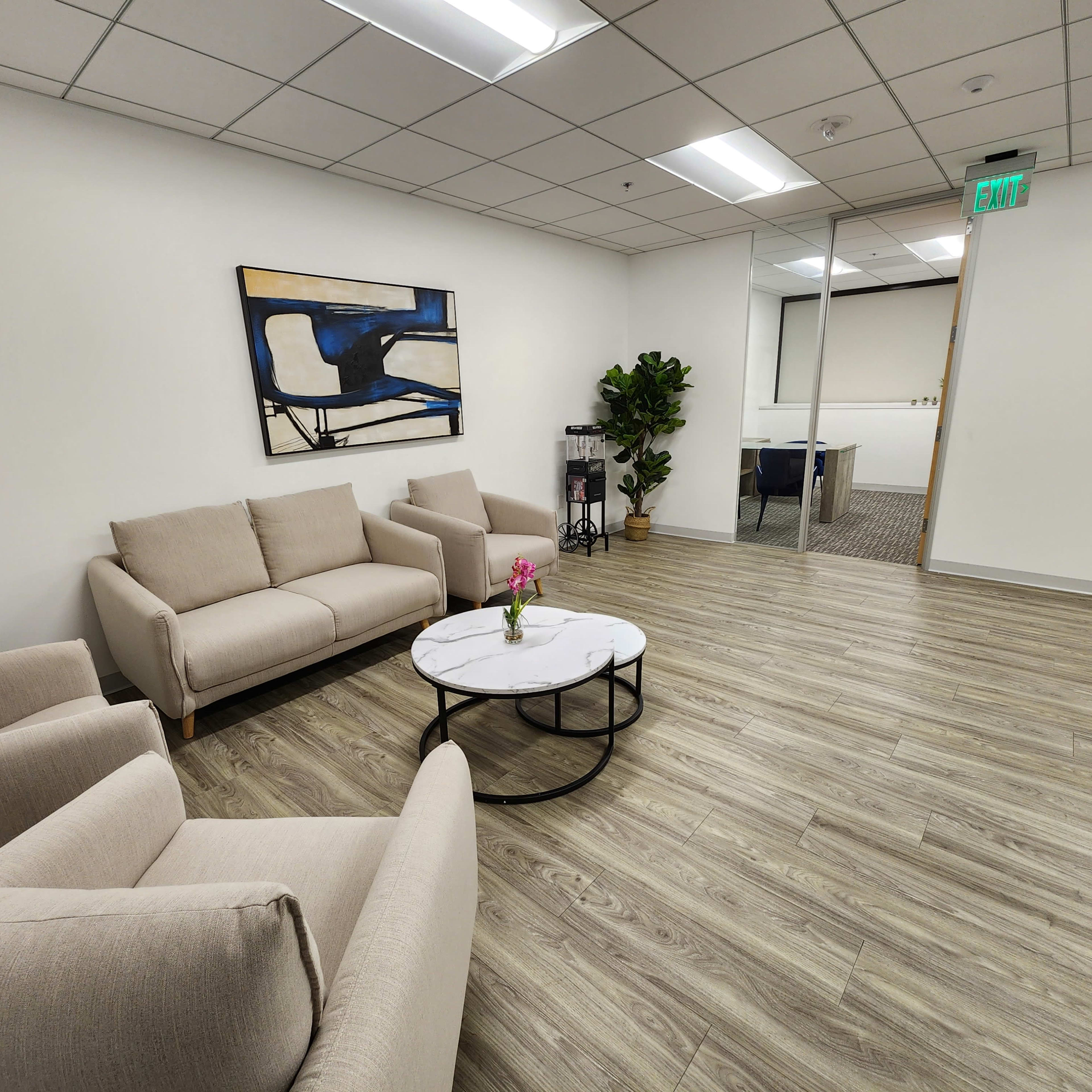 A modern waiting area with two light-colored sofas, a round coffee table, a decorative plant, and an abstract painting on the wall.
