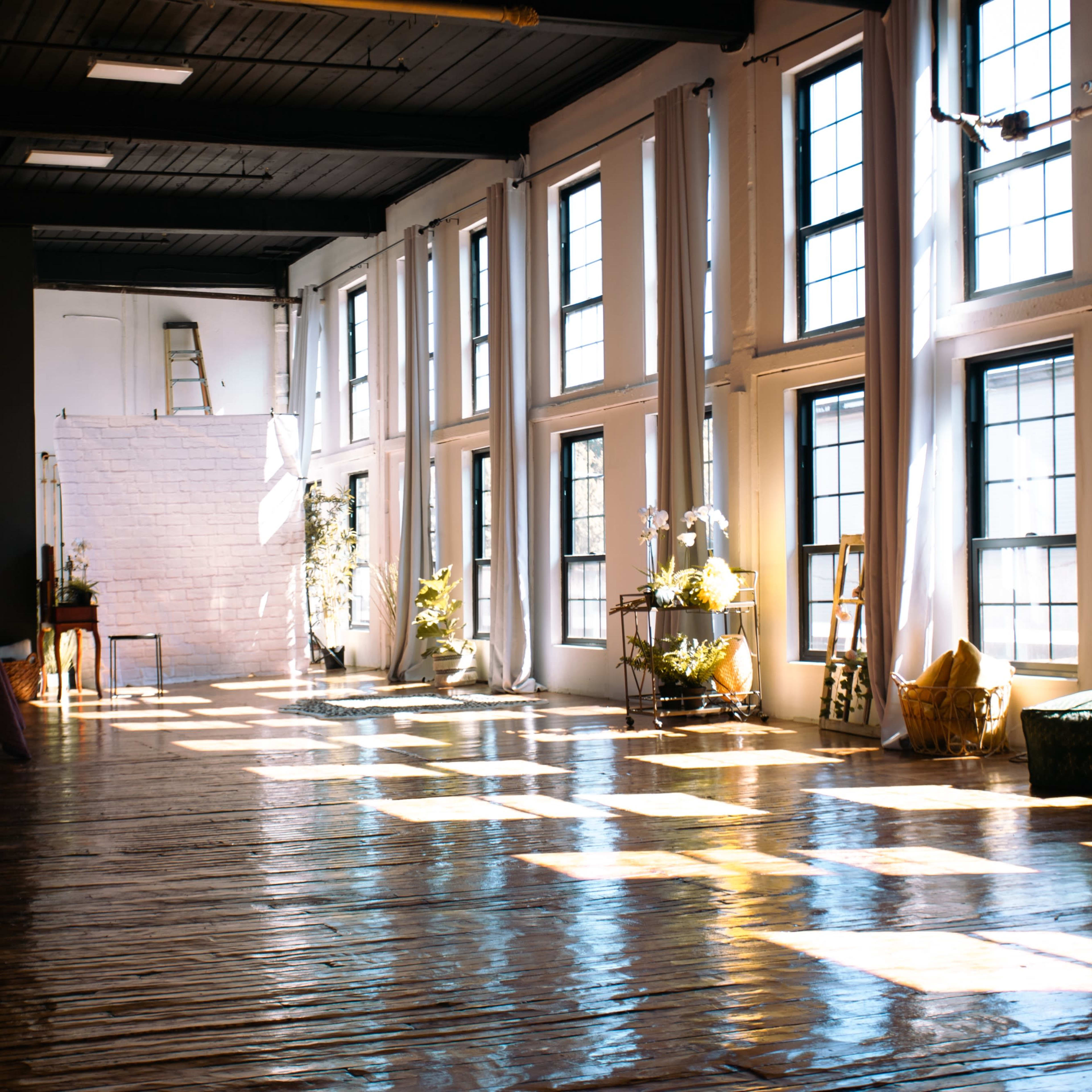 A spacious room features large windows that allow natural light to illuminate the wooden floor and various plants scattered throughout the space.