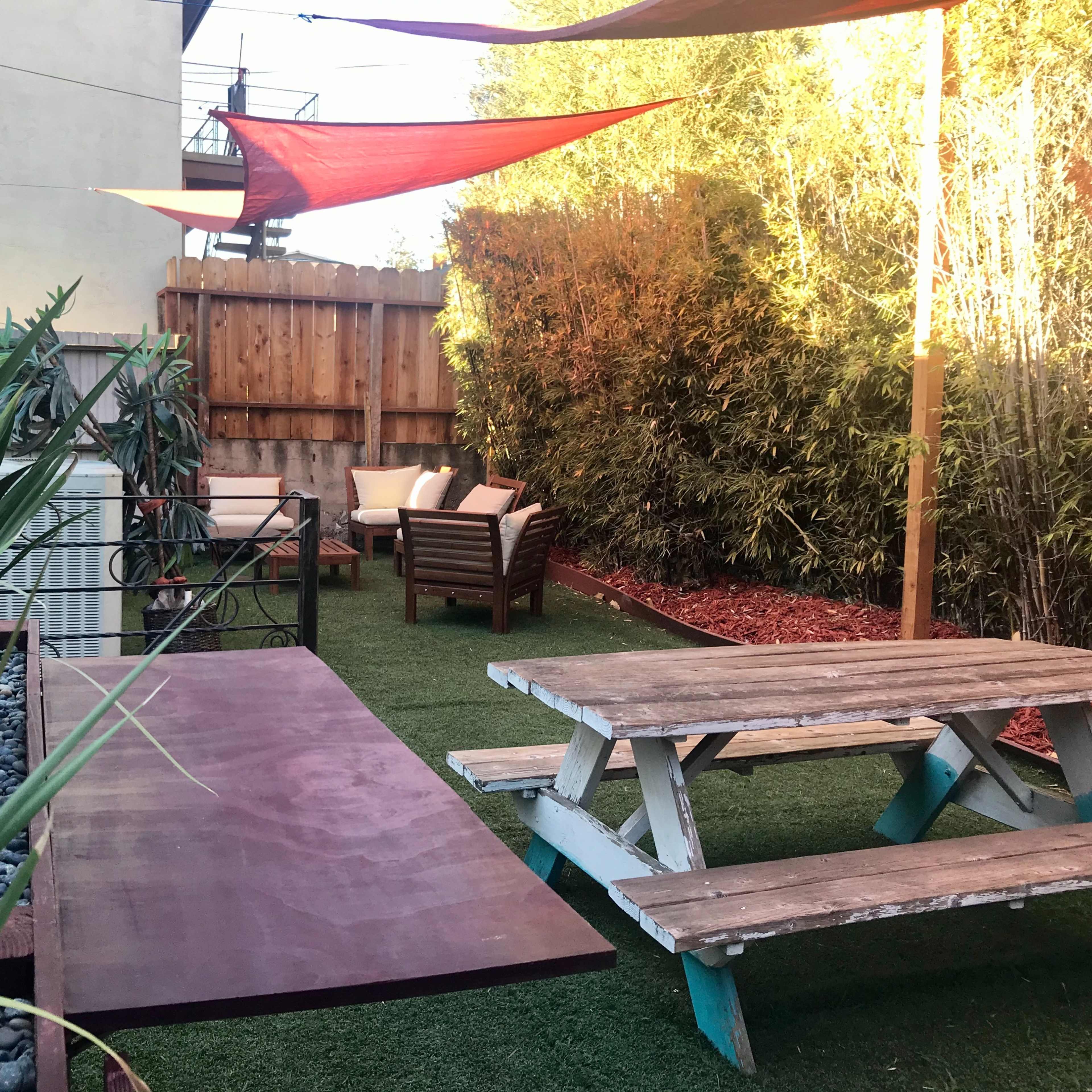 A backyard space with a picnic table, lounge chairs, and colorful shade sails above, surrounded by greenery and stones.
