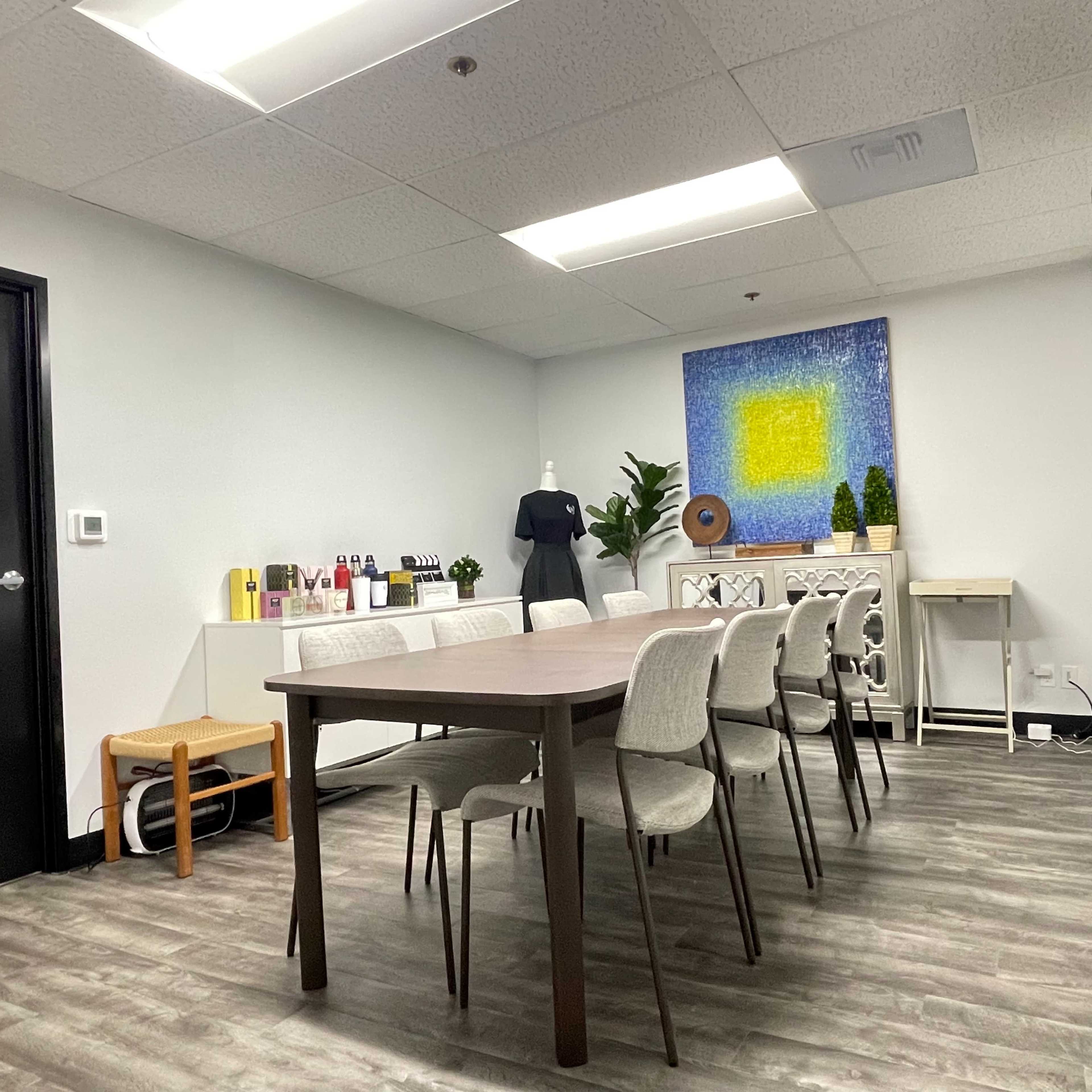 A conference room features a long table with eight chairs, a side table with decorative items, and a colorful wall art piece.