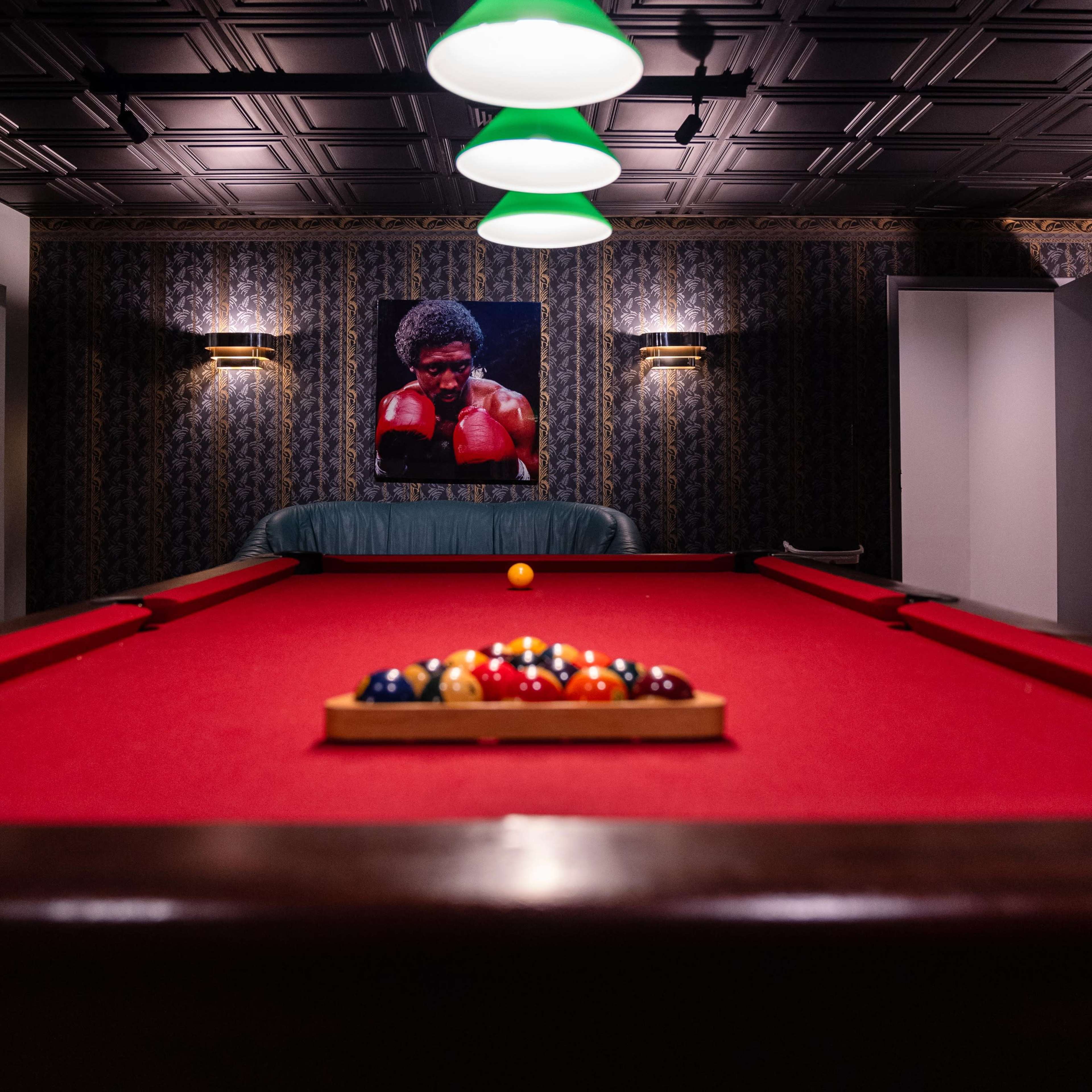 A billiards table with a set of balls arranged on it in a dimly lit room, featuring a large portrait of a boxer on the wall behind.
