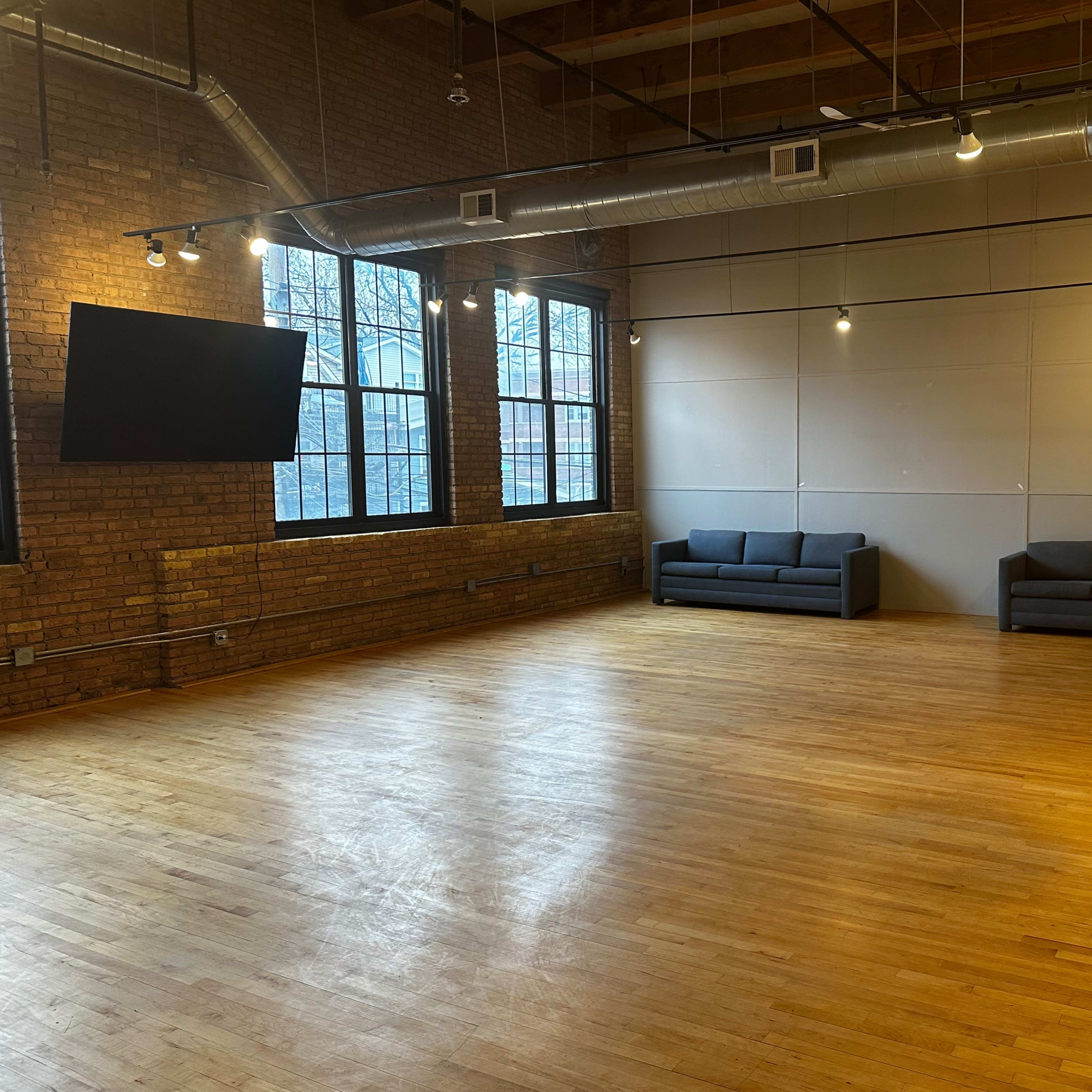 A spacious room with hardwood floors, featuring two gray sofas against a brick wall, and large windows that provide natural light.