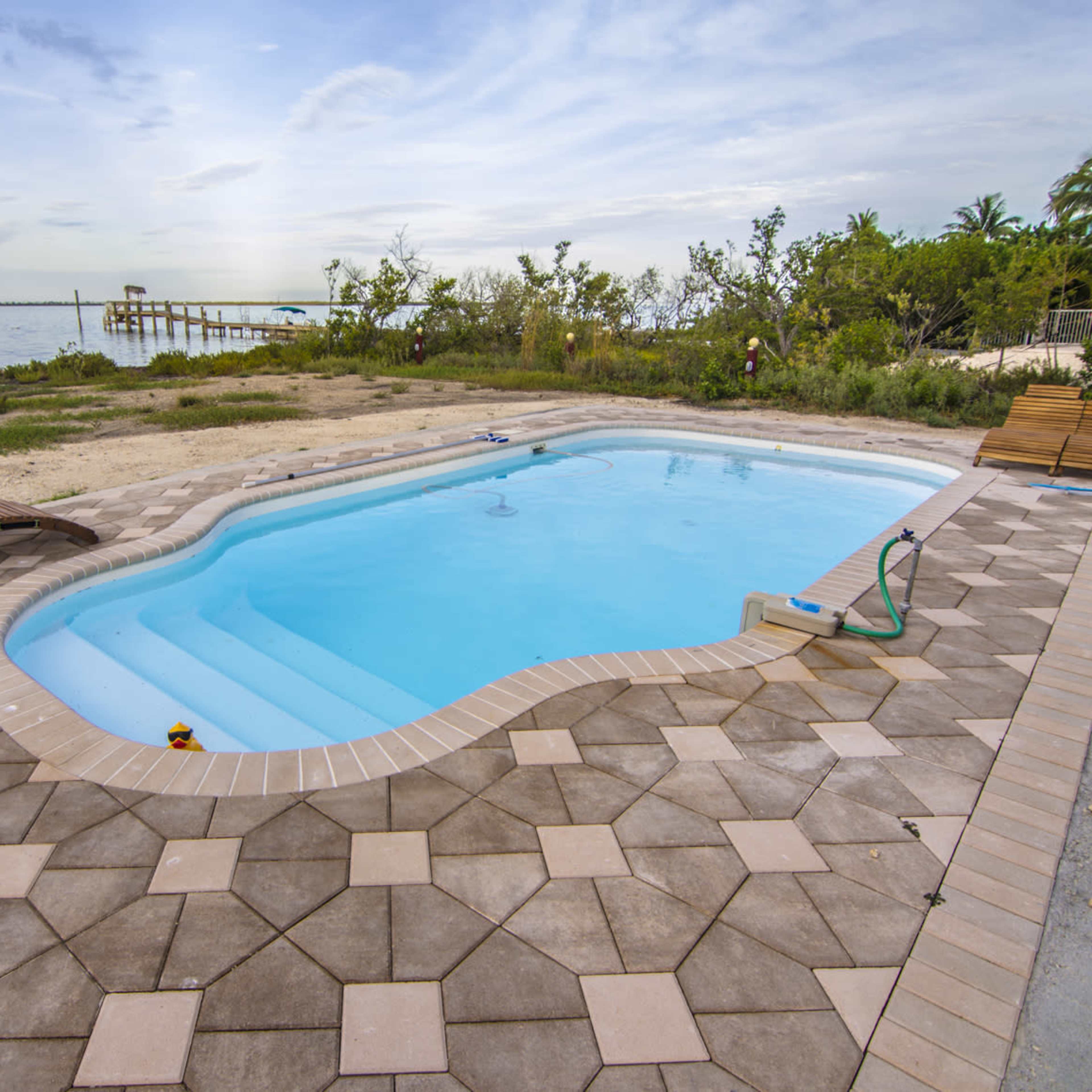 A swimming pool with a light blue surface is surrounded by a tiled patio and overlooks a calm body of water, with a wooden deck and vegetation in the background.
